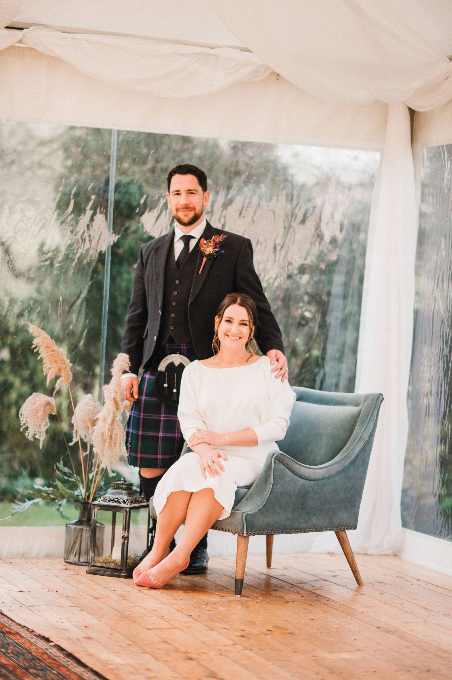 Wedding couple poses by modern gray chair in a white tent with pampas grass decor on wooden floor.