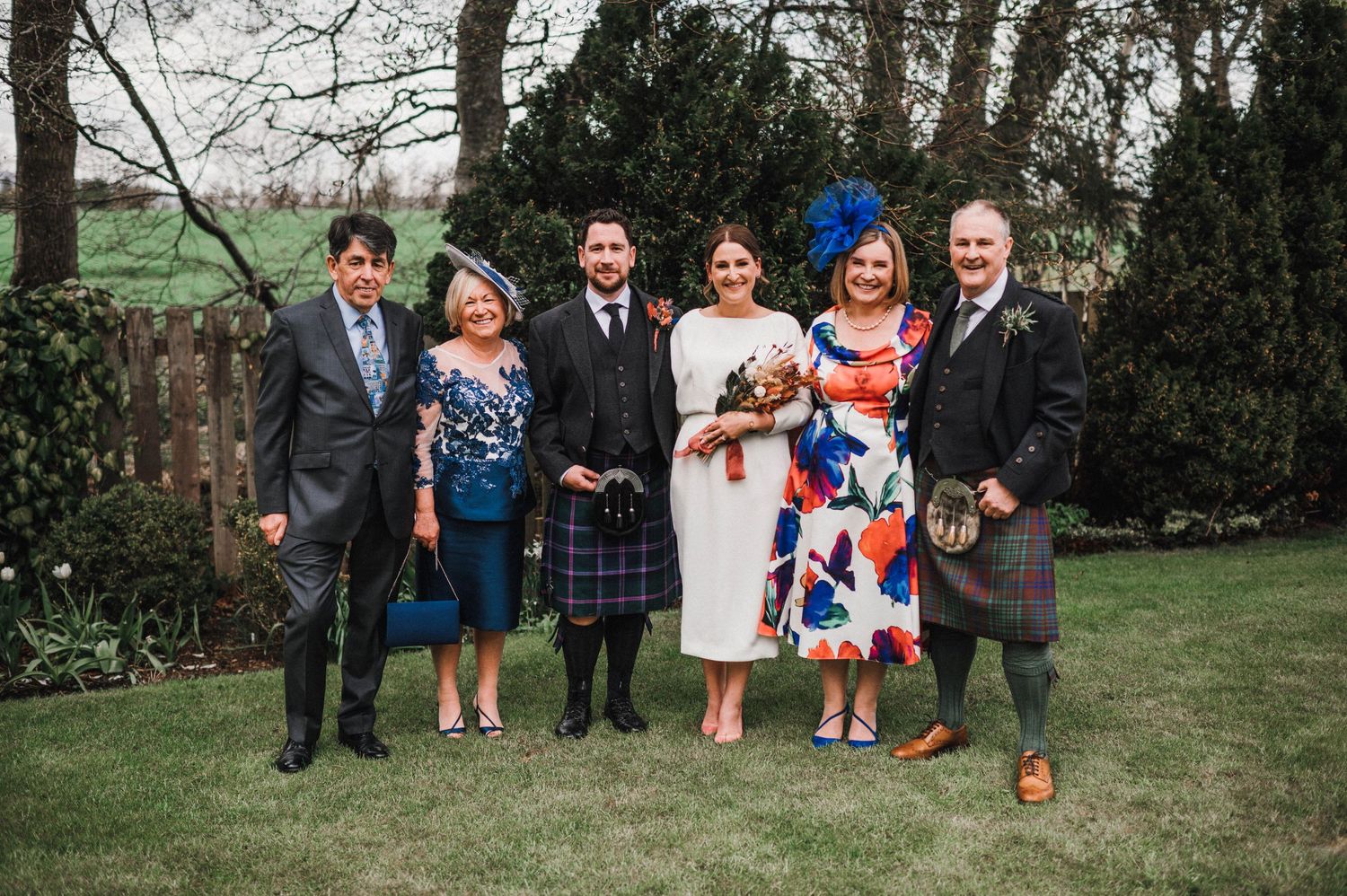 Wedding party poses together outdoors wearing mix of kilts and formal attire with Scottish elements.