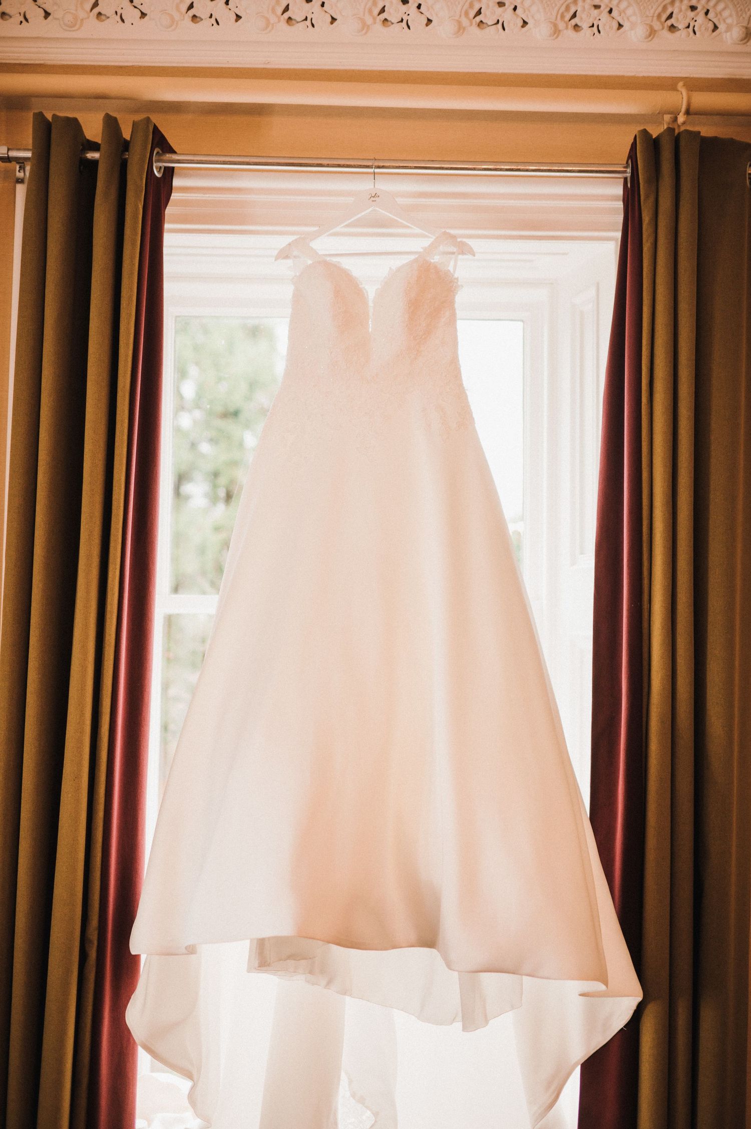 A classic white wedding gown displayed against natural window light and draped curtains.