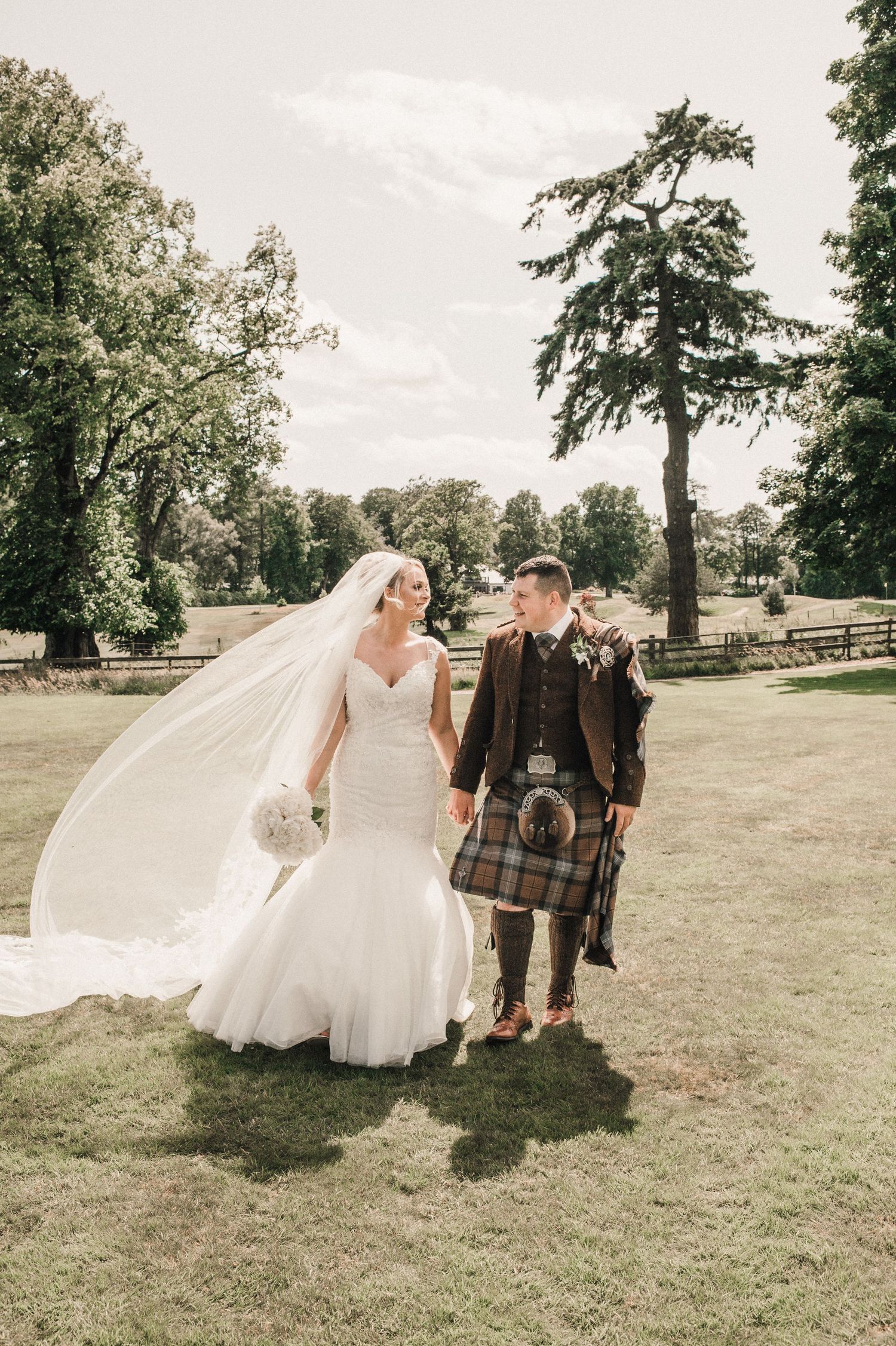 Wedding couple walks hand in hand on grass with bride in white gown and groom in traditional kilt.