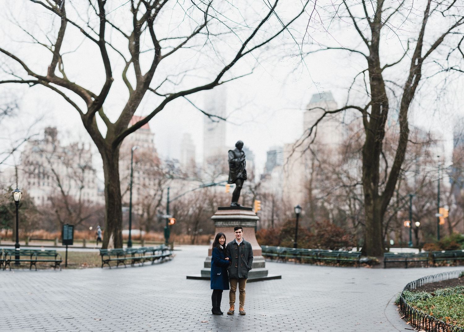 A couple poses near a statue in Central Park with winter trees and city buildings in the background.