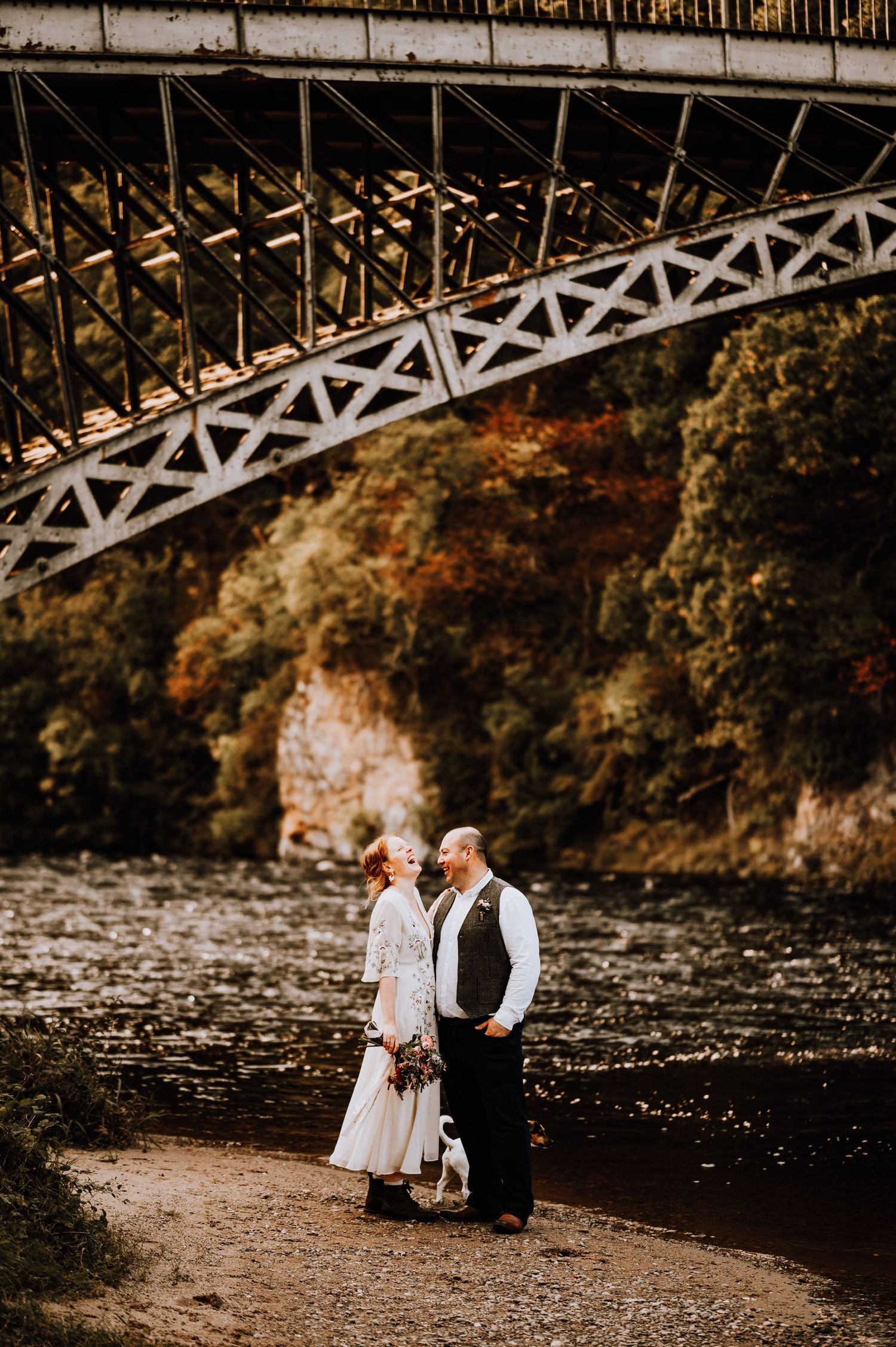 A couple stands beneath a dramatic iron bridge structure overlooking a flowing river.