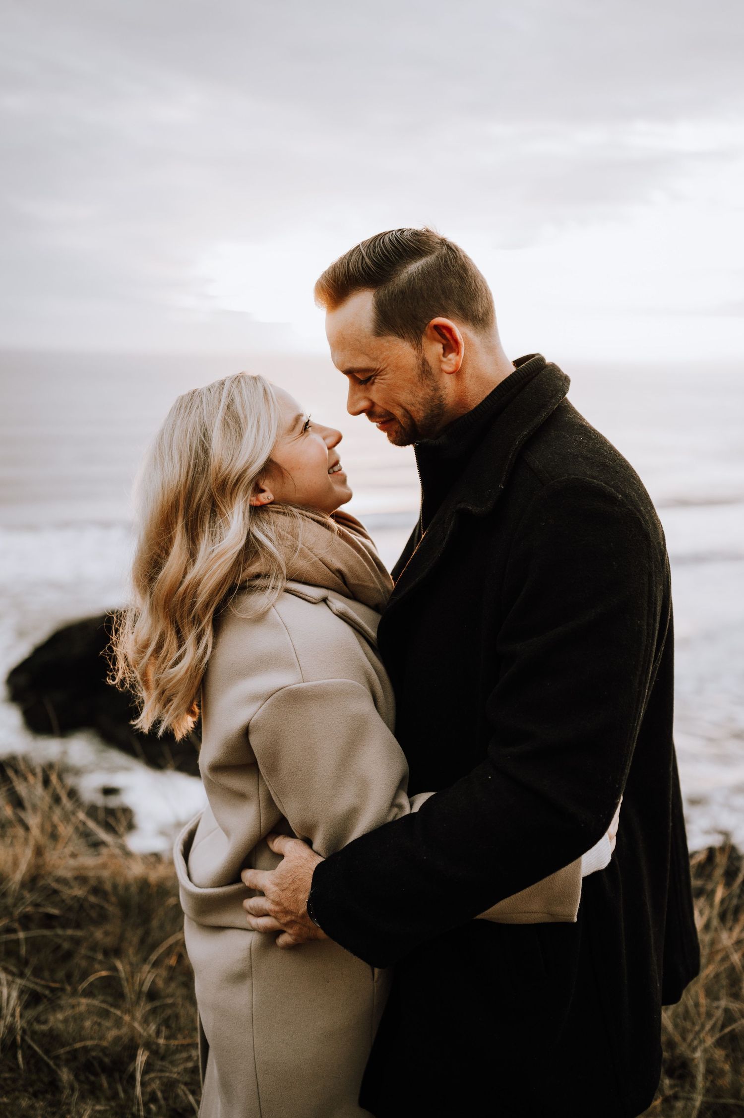 Couple in elegant winter coats sharing a tender moment against a coastal backdrop.