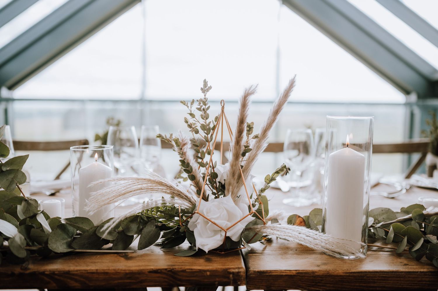 Elegant wedding centerpiece with dried flowers and white candles on a wooden table in a glass-roofed venue.