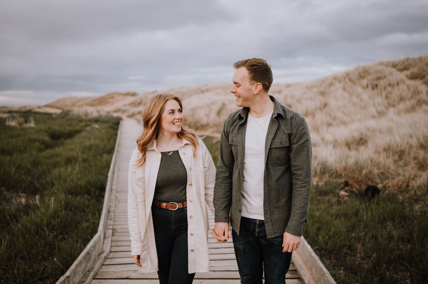 A couple walks together on a wooden boardwalk through coastal dunes.