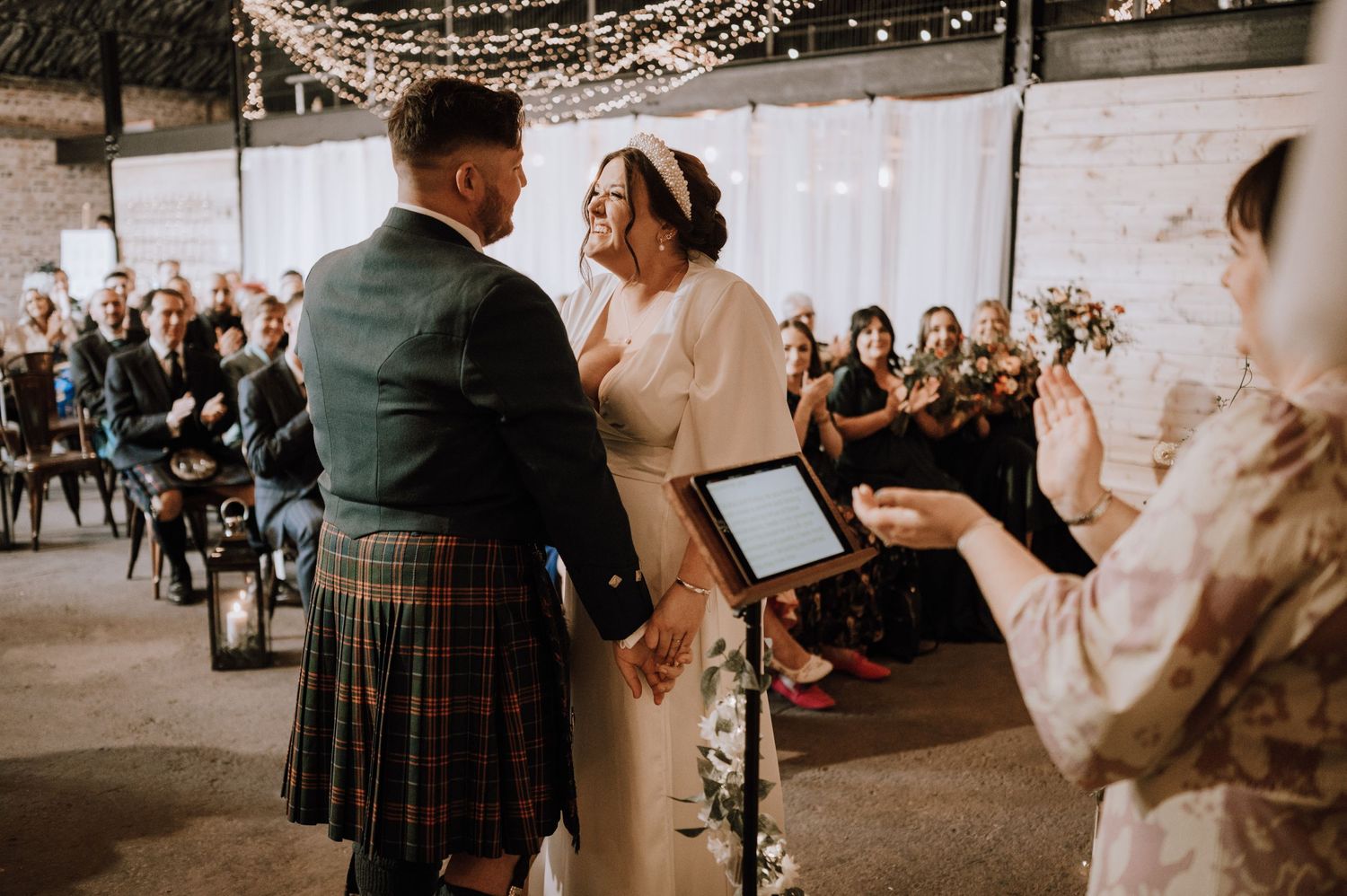 Wedding ceremony moment in industrial venue with string lights and gathered guests.