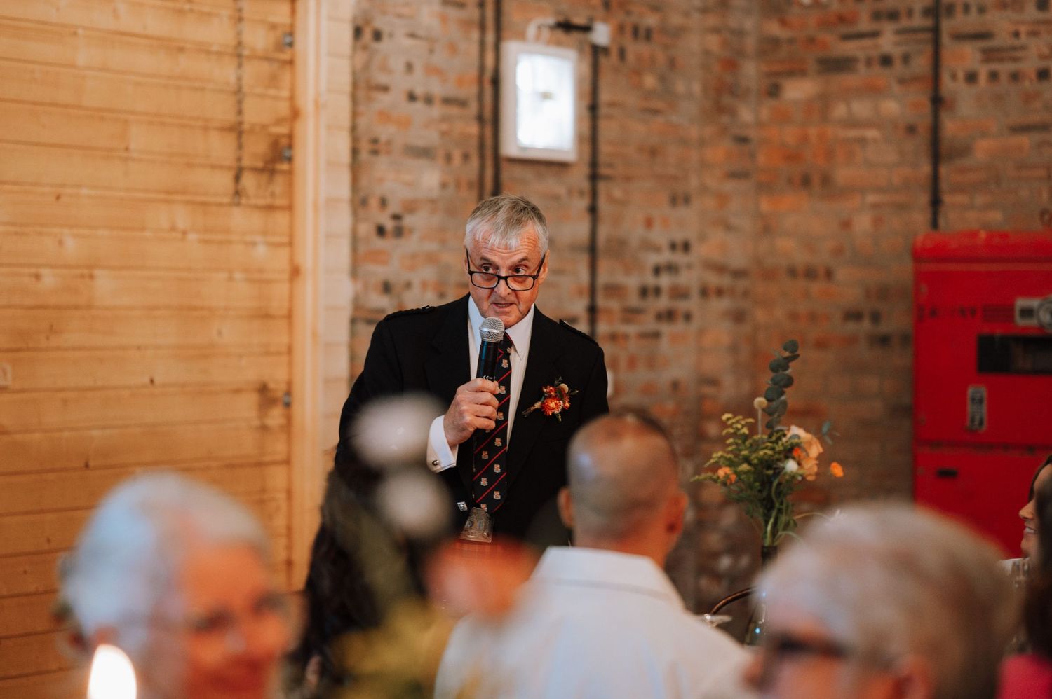 Guest speaking at wedding reception in rustic brick-walled venue.