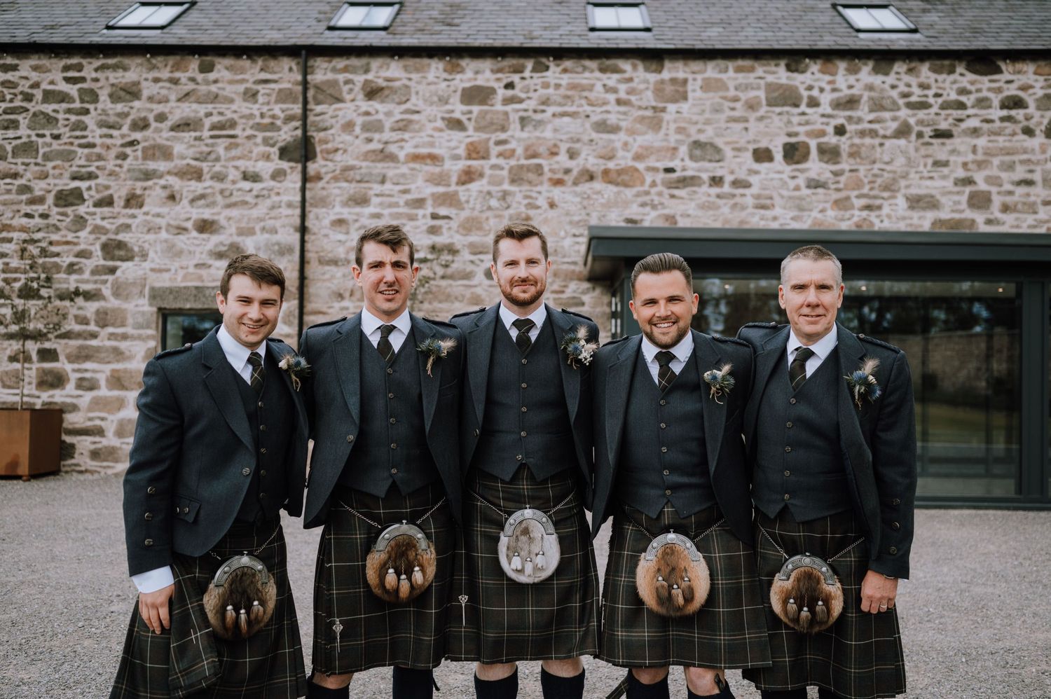 Five men in traditional Scottish kilts and formal jackets pose outside a stone building.