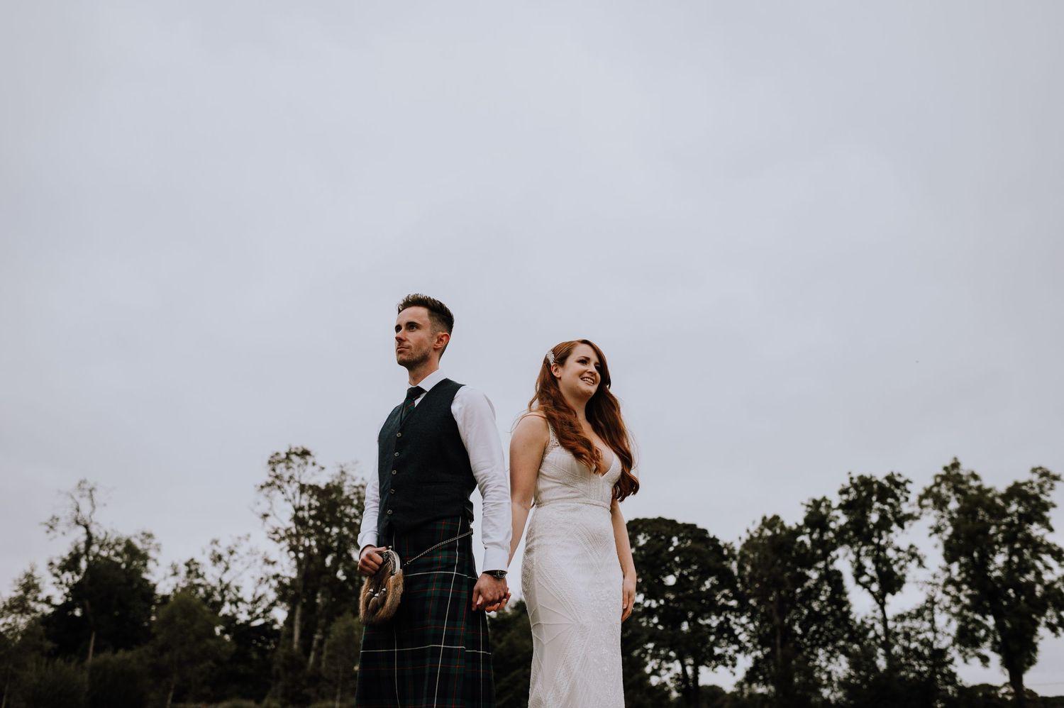 Bride and groom stand dramatically against grey sky with trees in moody Scottish wedding photo.