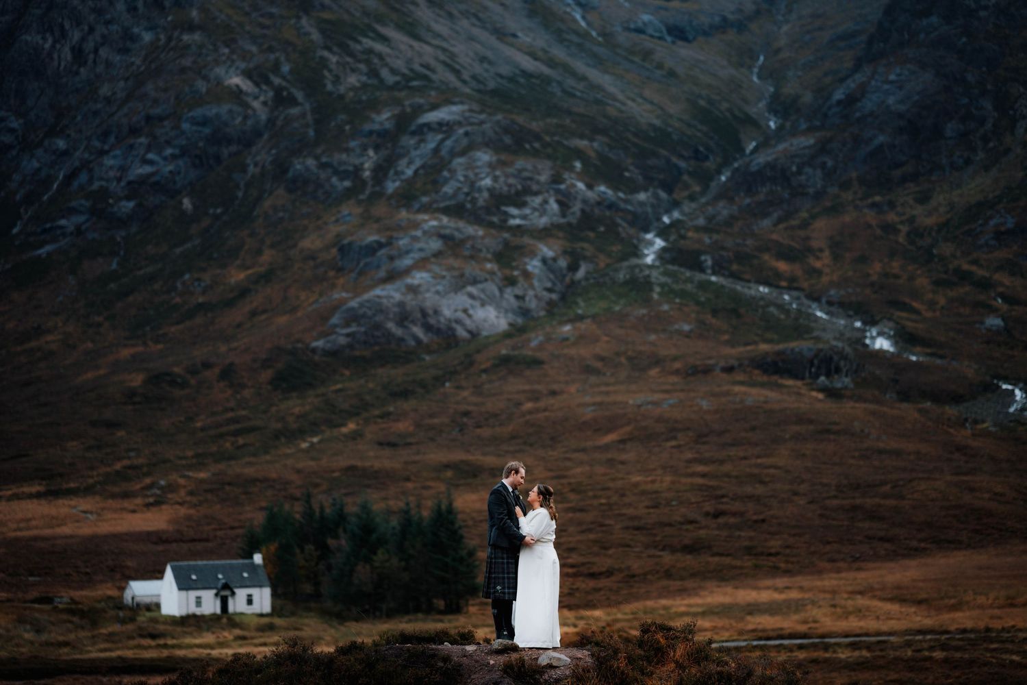 A wedding couple poses against dramatic mountain landscape with white cottage in Scottish Highlands.