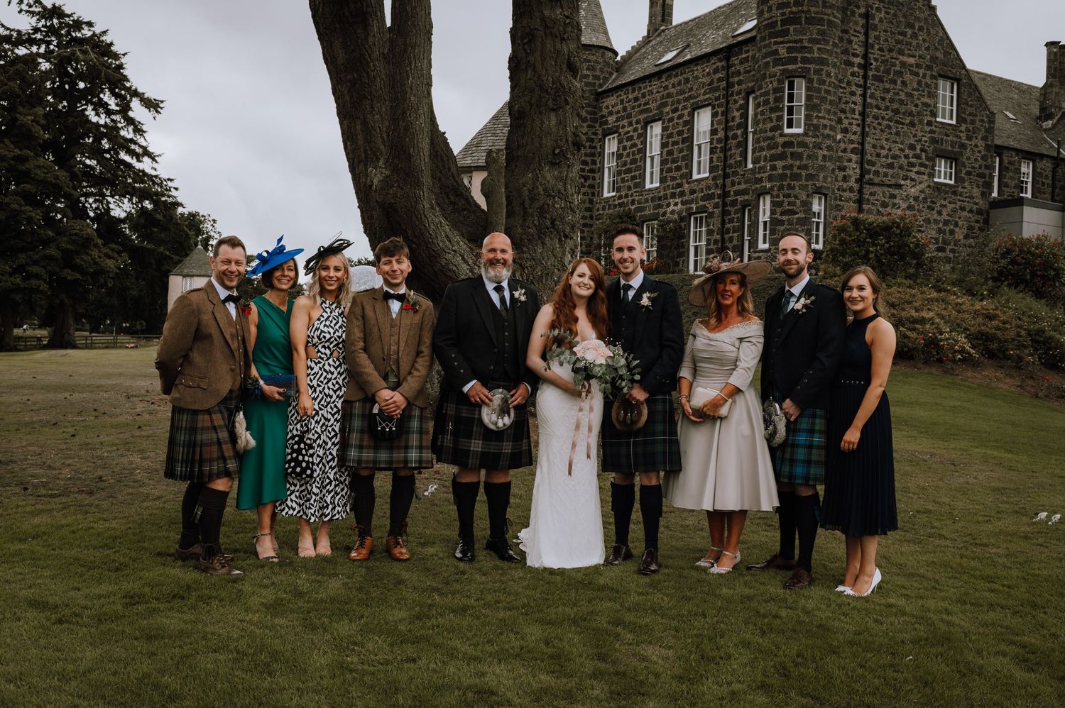 Group photo of wedding party wearing traditional Scottish kilts and formal attire by stone castle.
