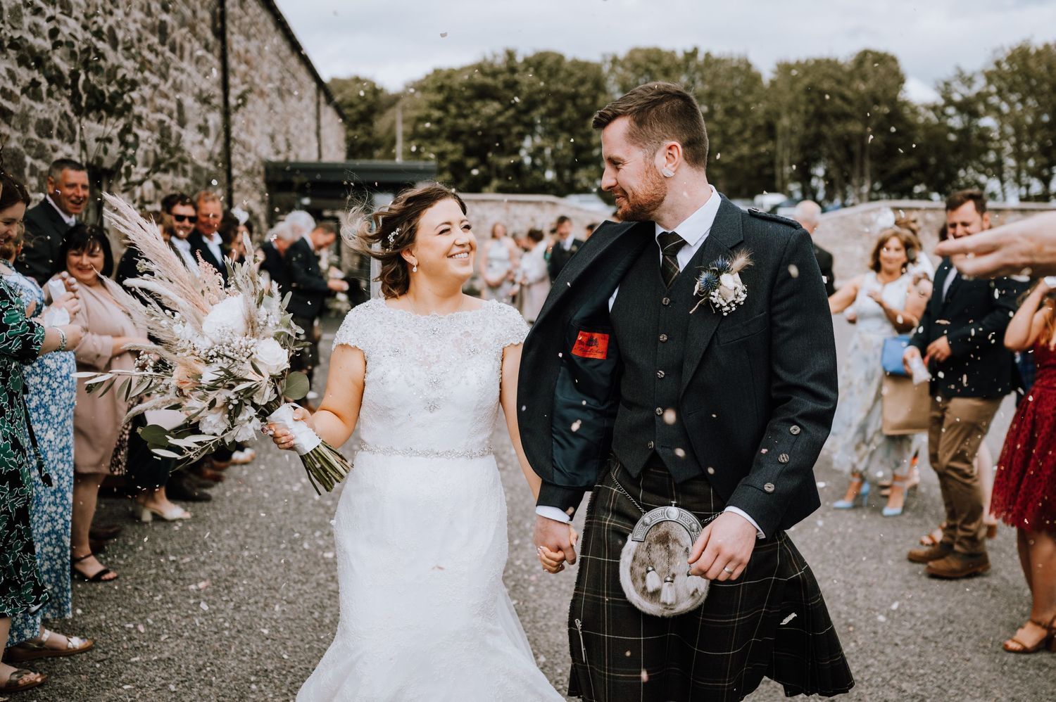 Wedding guests shower confetti over newlyweds outside stone building as groom wears traditional Scottish kilt.