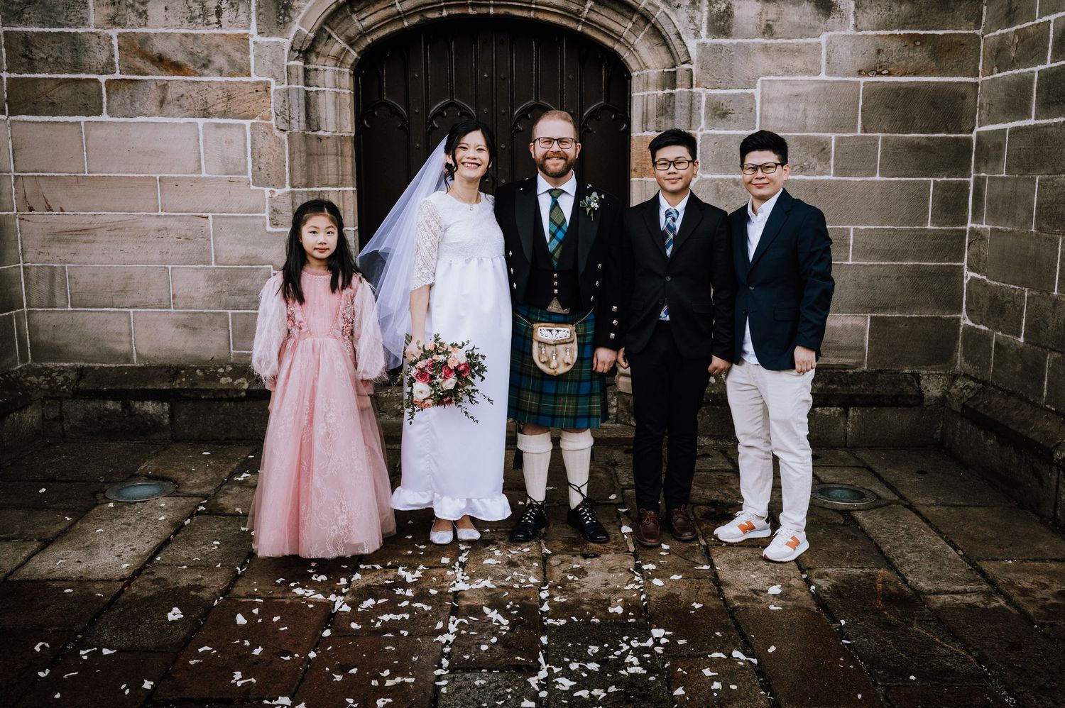 Wedding party group photo at stone church entrance with flower petals scattered.