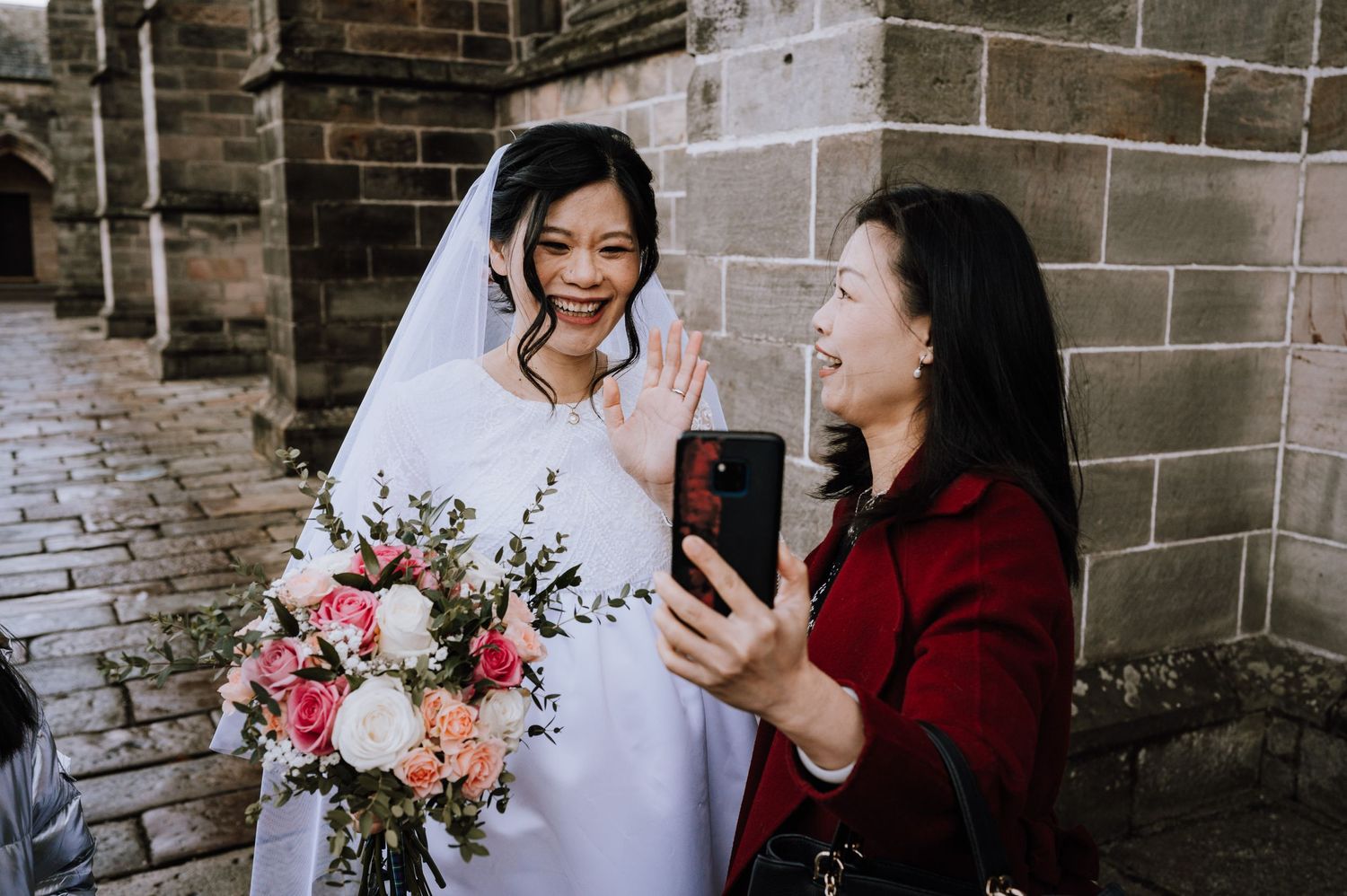A bride wearing a white dress holds a pink and white bouquet while someone takes a selfie with her.