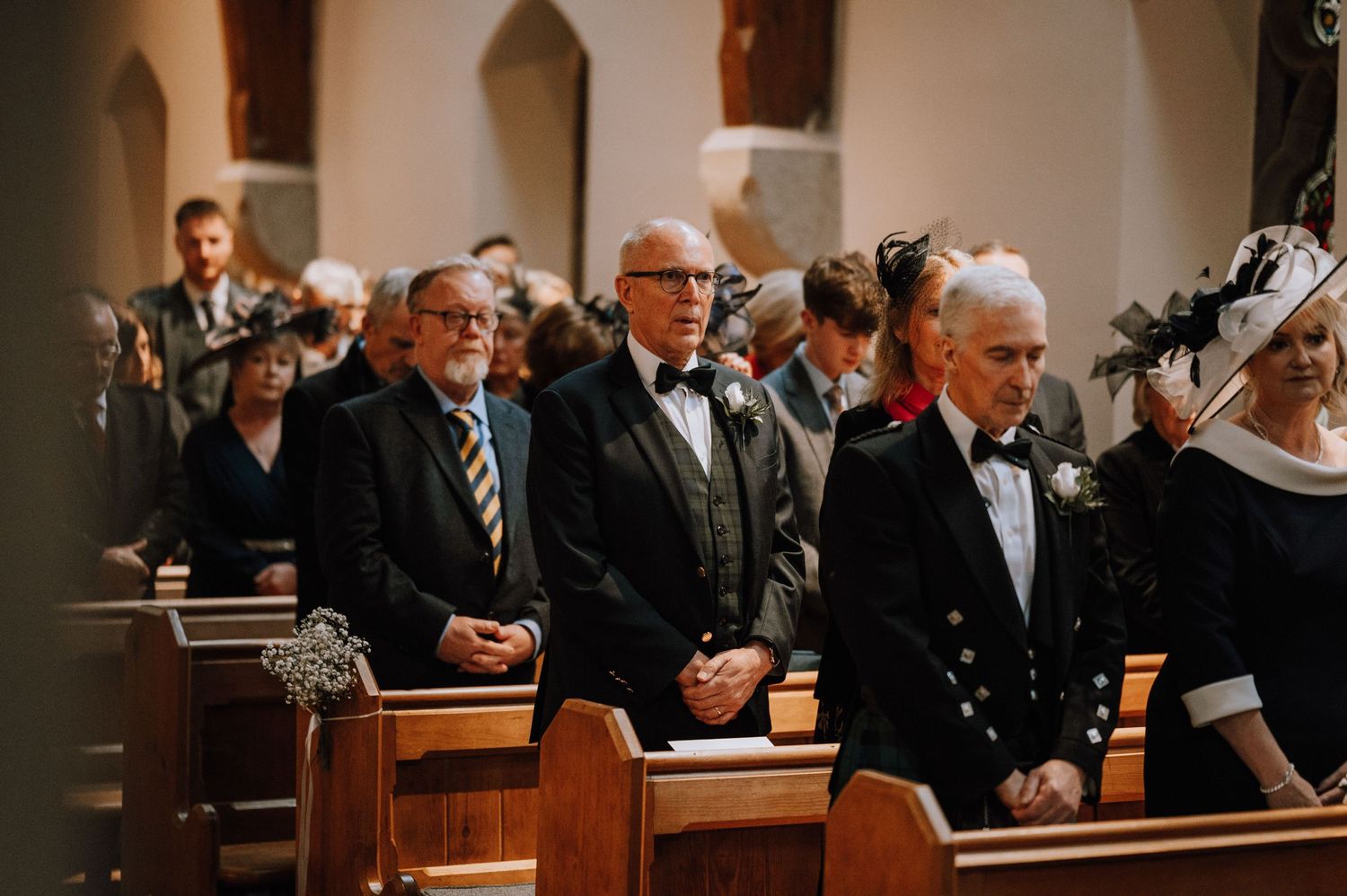 Wedding guests in formal attire stand in church pews during a traditional ceremony.
