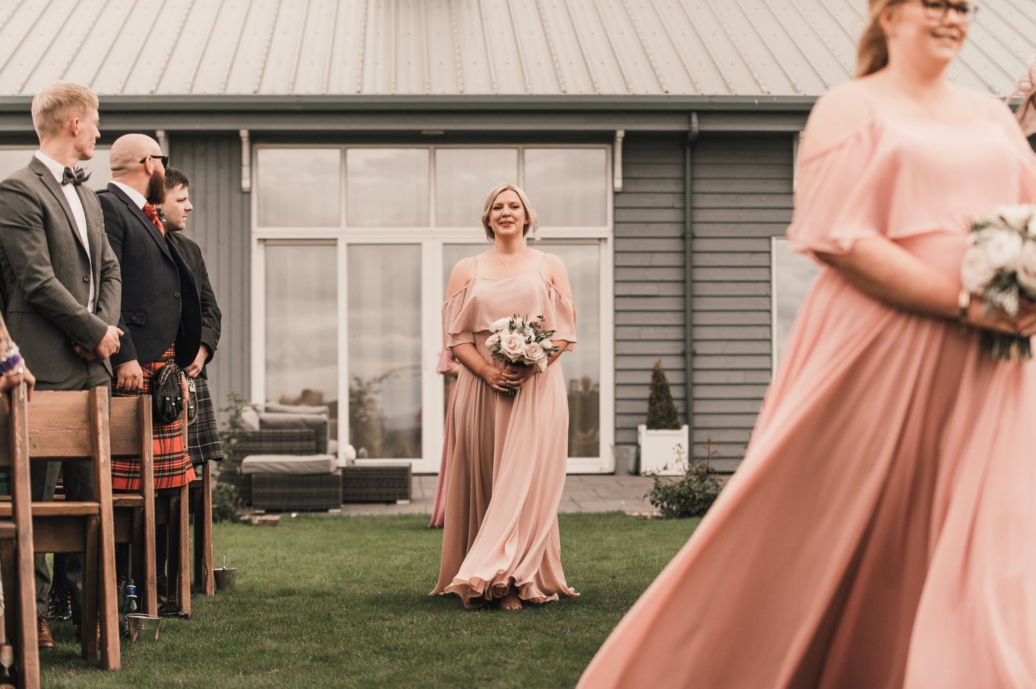 Wedding ceremony setup with bridesmaids in blush dresses walking down an outdoor aisle.