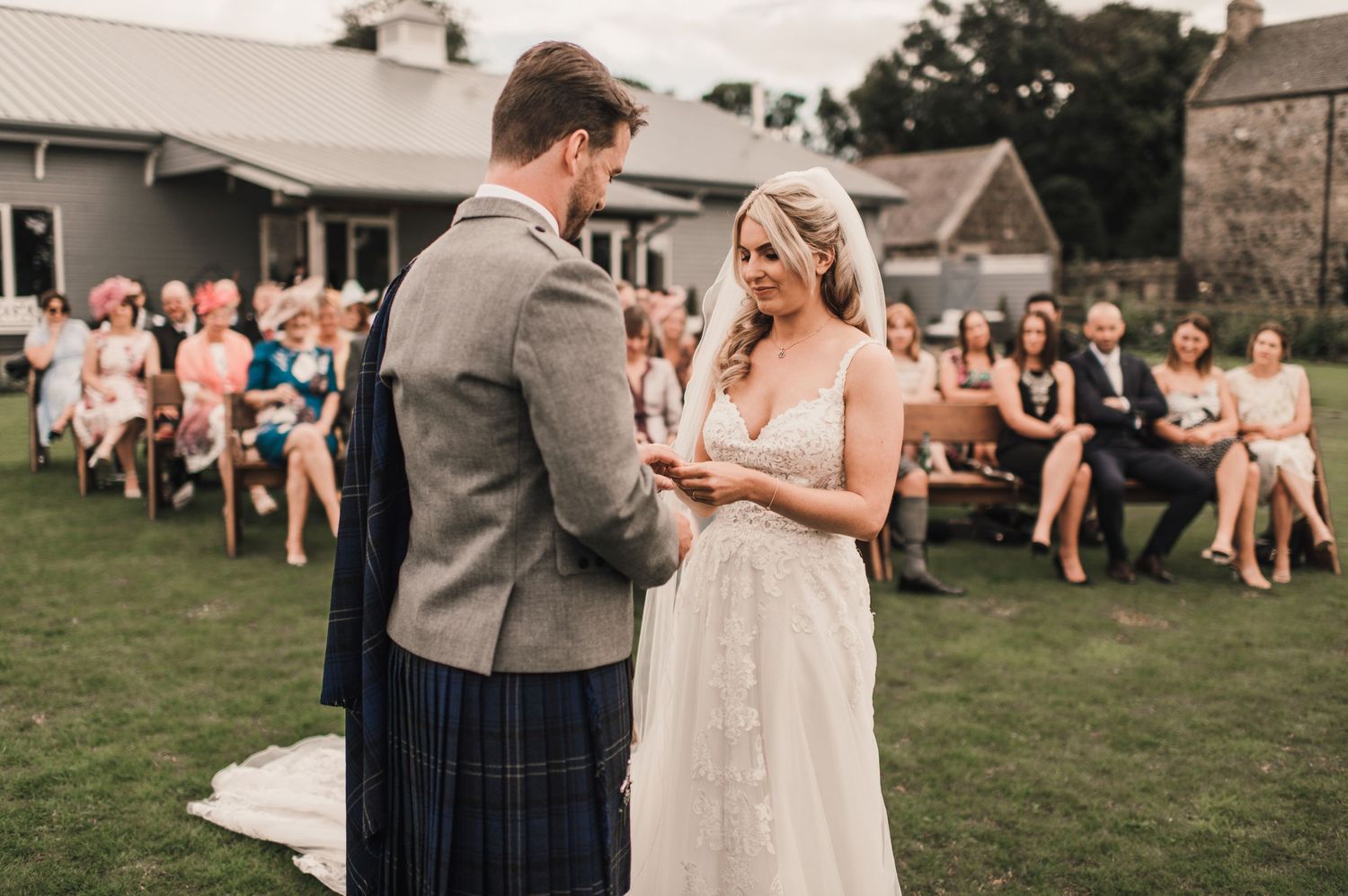A couple exchanges rings during their outdoor wedding ceremony while guests watch from chairs.