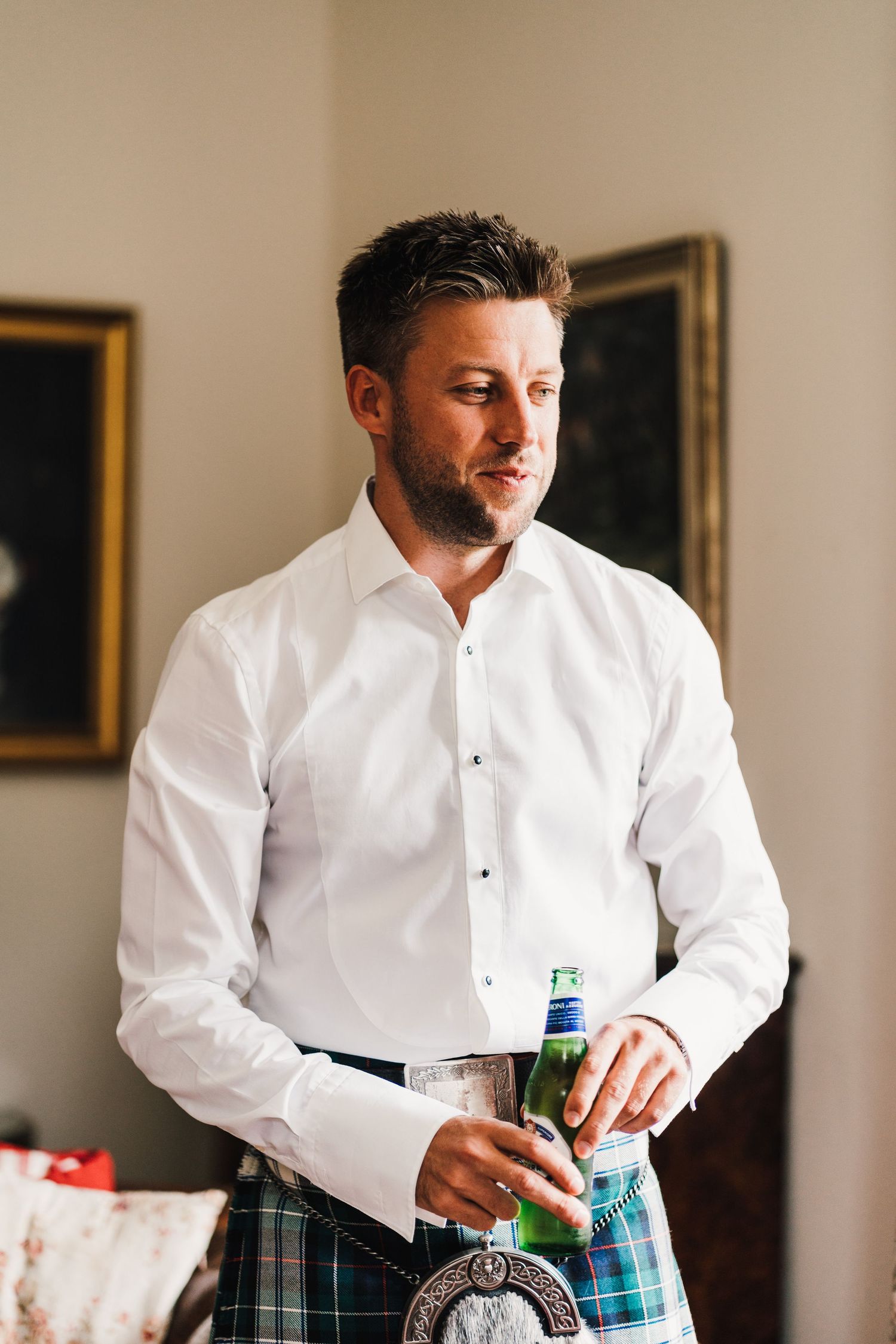 Man in white dress shirt and traditional Scottish kilt holds a beverage.