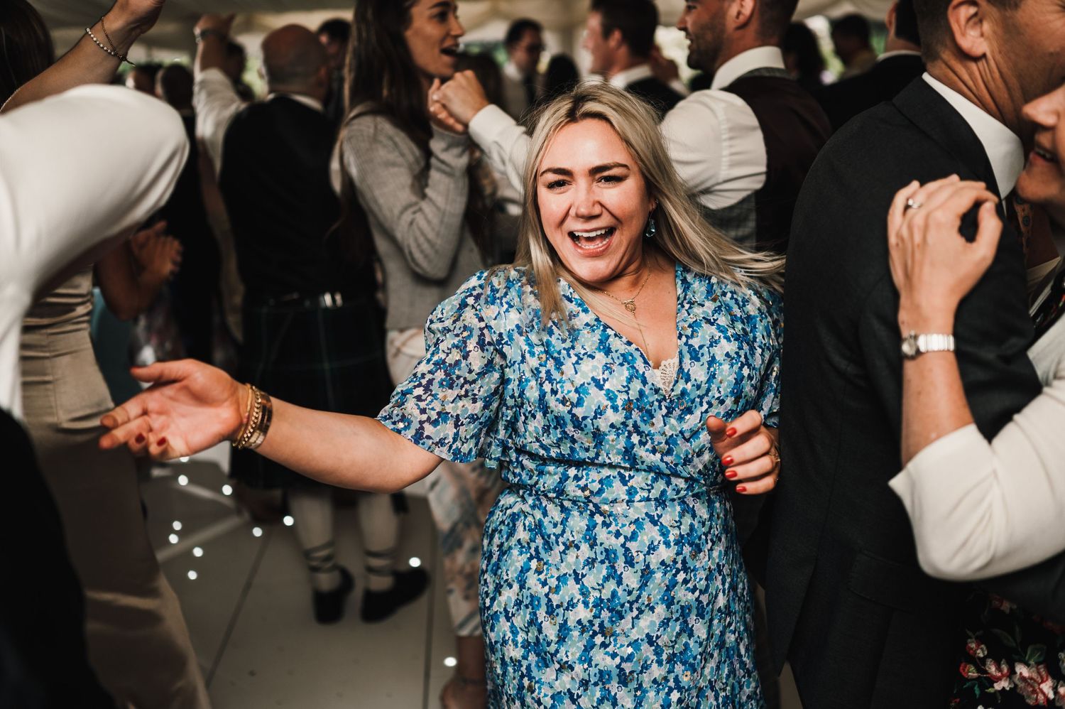 Wedding guest in blue floral dress dancing enthusiastically at reception party.