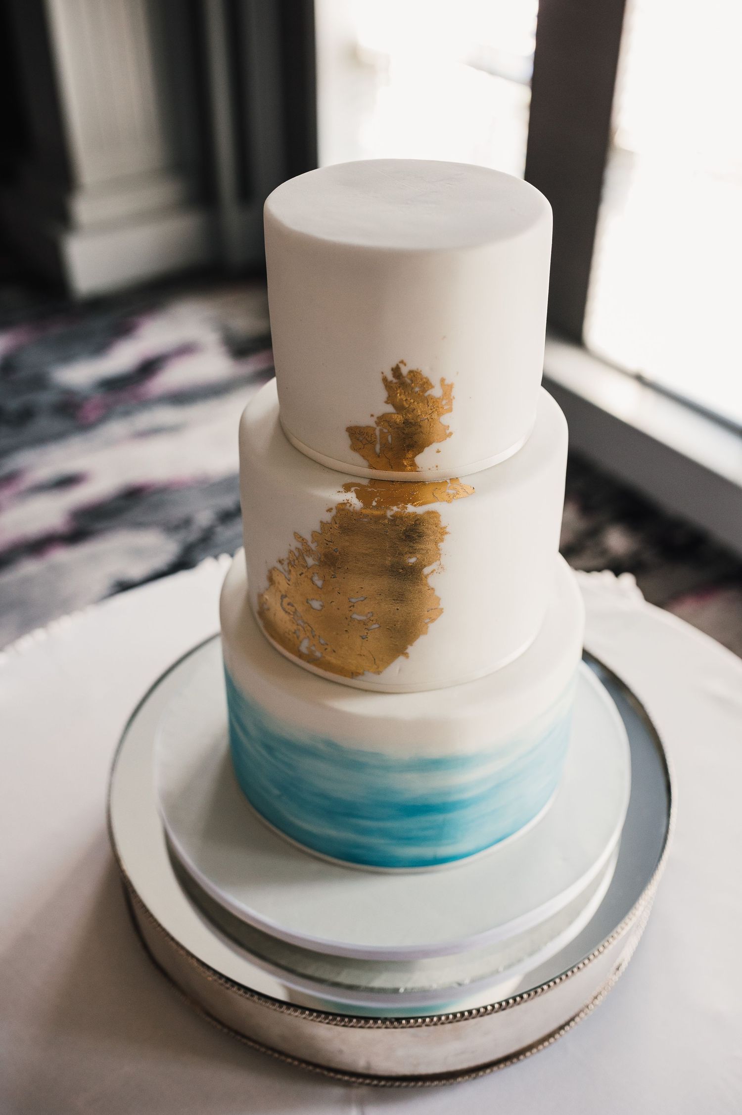 Elegant three-tier wedding cake featuring white frosting, gold accents, and watercolor blue base design.