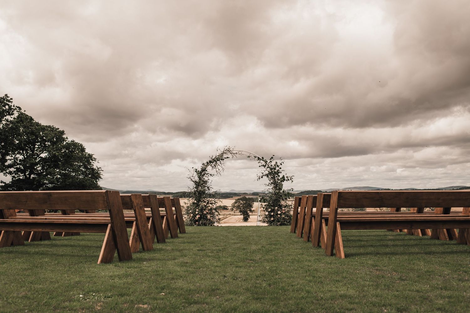 Wooden benches line an outdoor wedding ceremony aisle leading to a floral arch under dramatic cloudy skies.