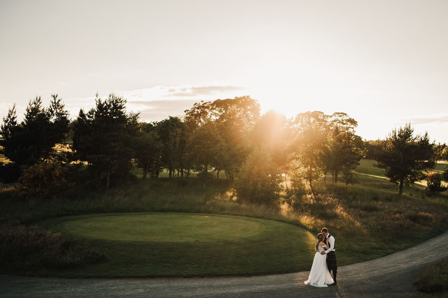 A small silhouetted couple stands on a golf course path during a dramatic golden sunset.