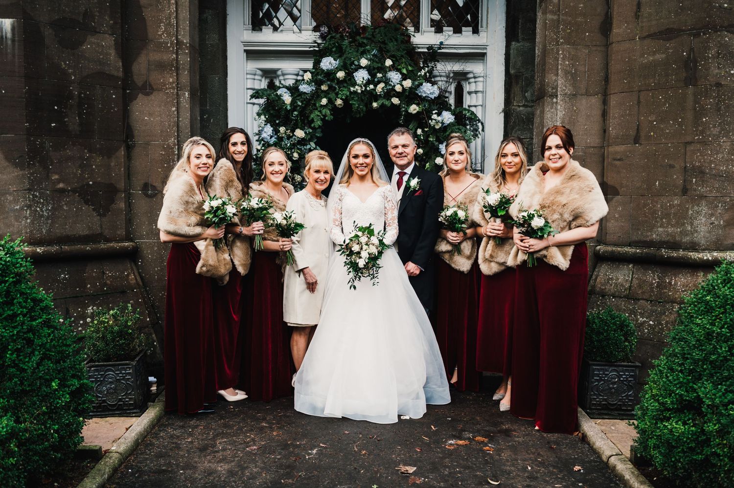 Wedding party in burgundy dresses and beige shawls pose outside a stone church entrance.