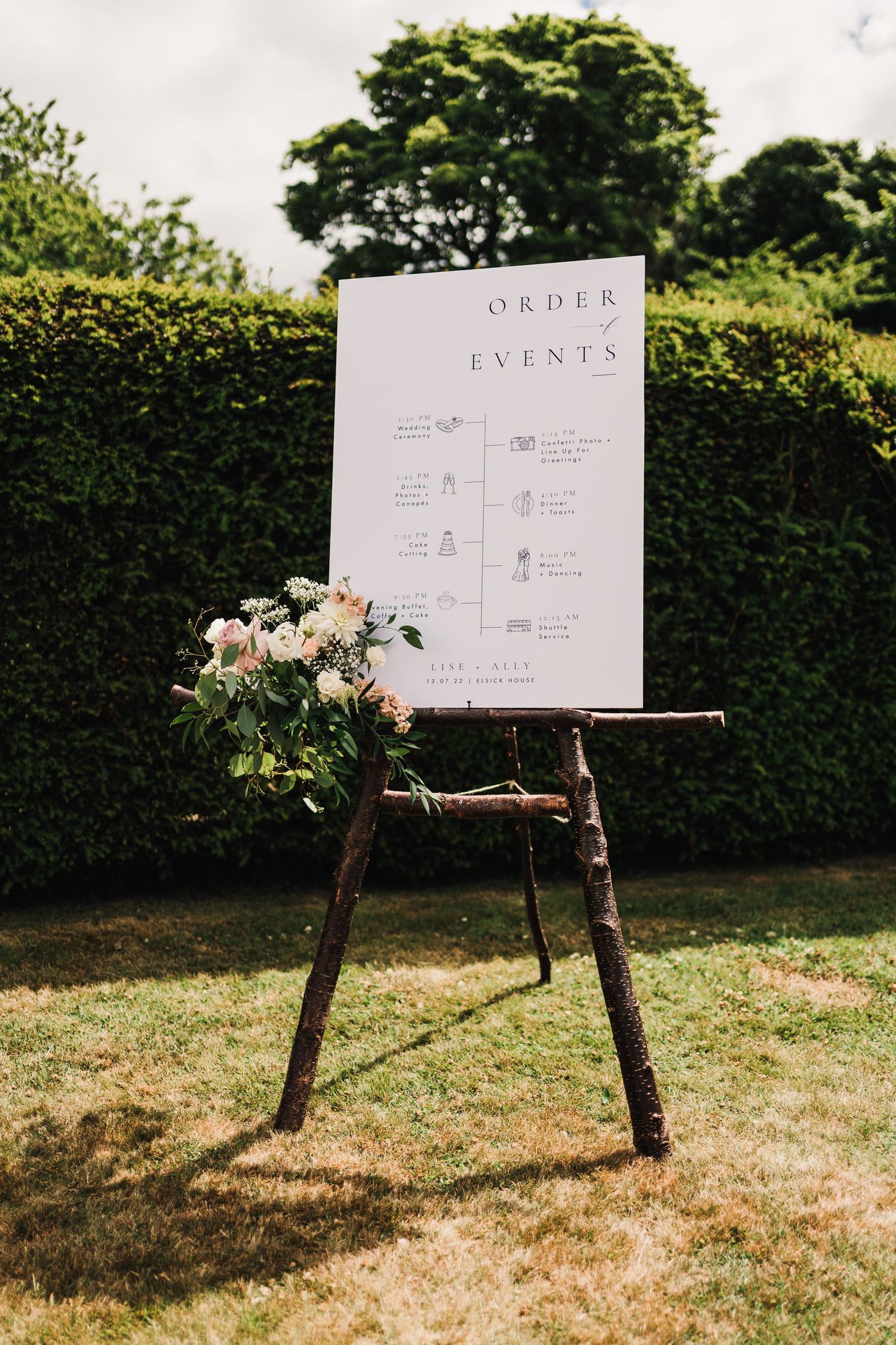 Wedding event schedule displayed on white board with rustic wooden easel and floral arrangement.