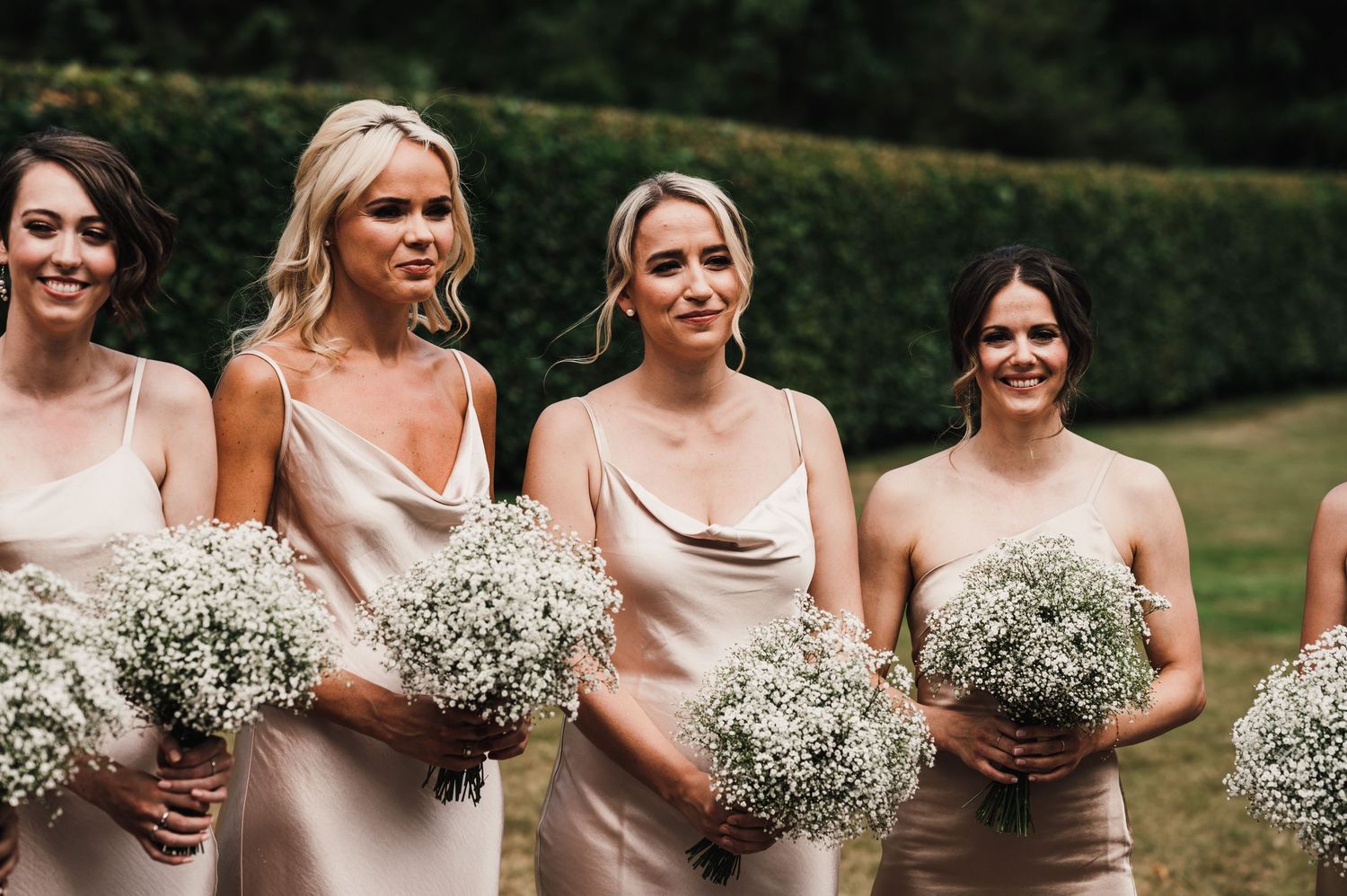 Bridesmaids in blush silk dresses holding baby's breath bouquets outdoors.