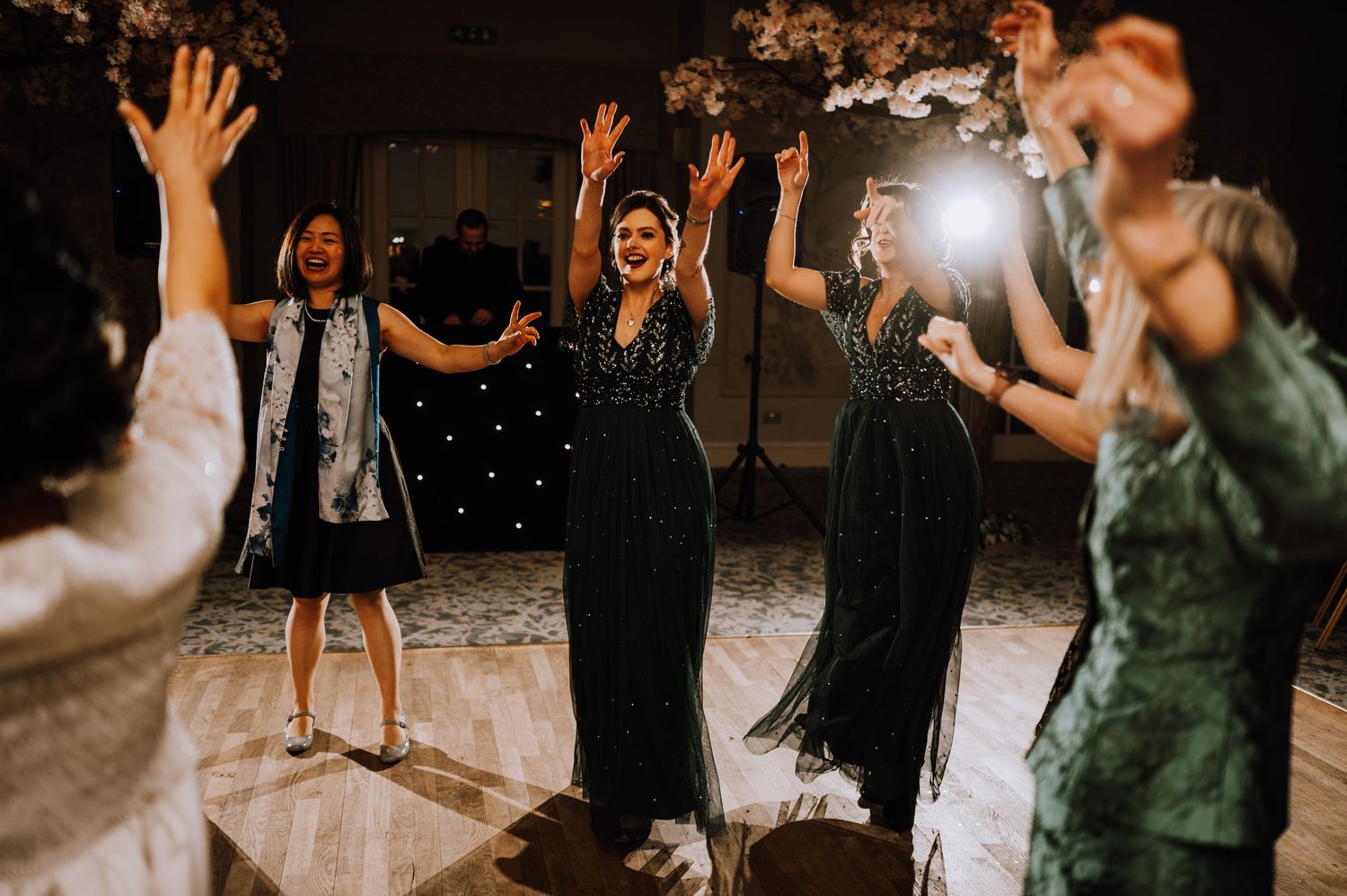 Guests raise their hands while dancing at a dimly lit wedding reception.