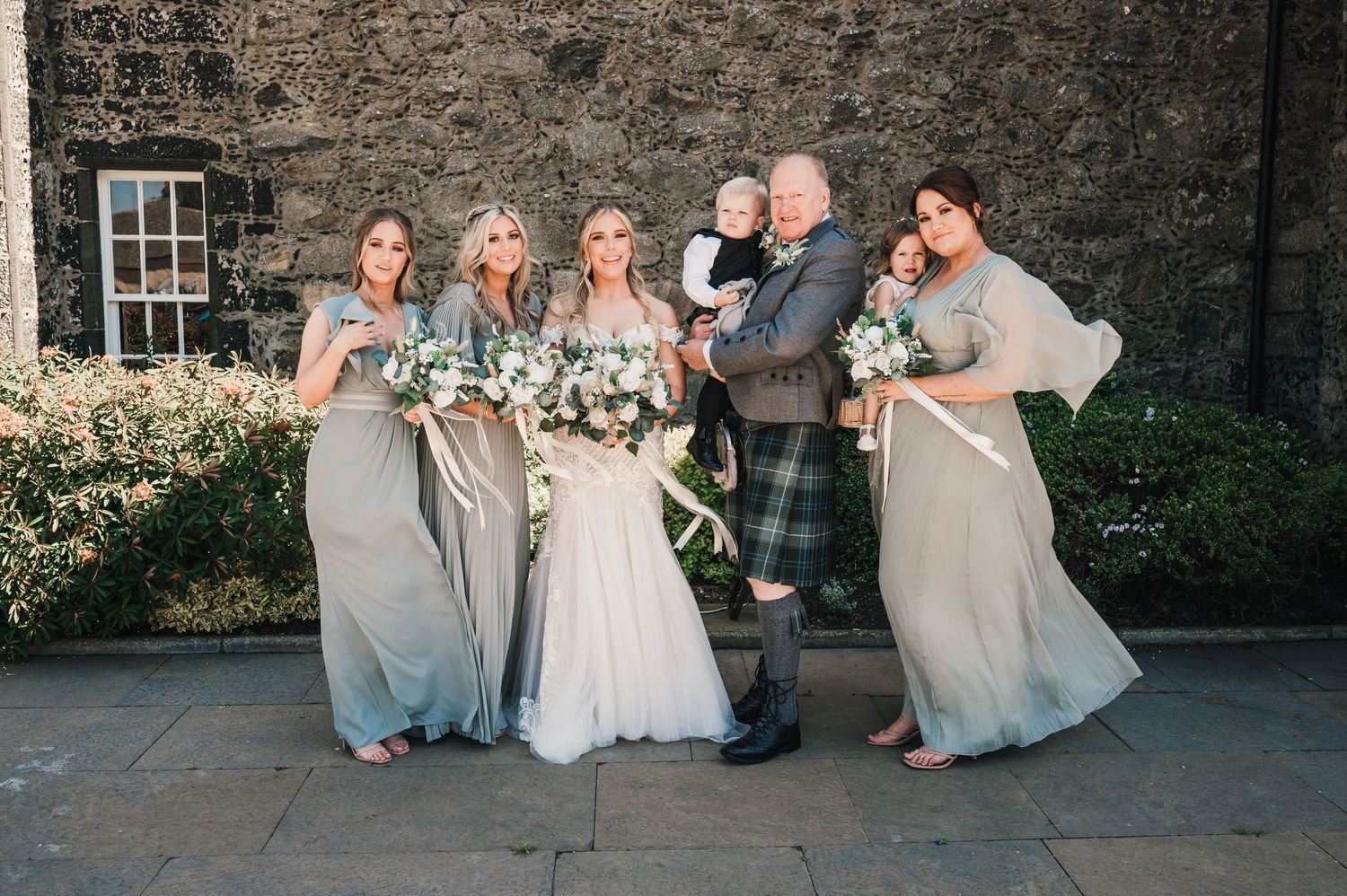 Wedding party poses for fun photo outside stone building with bridesmaids in grey dresses.