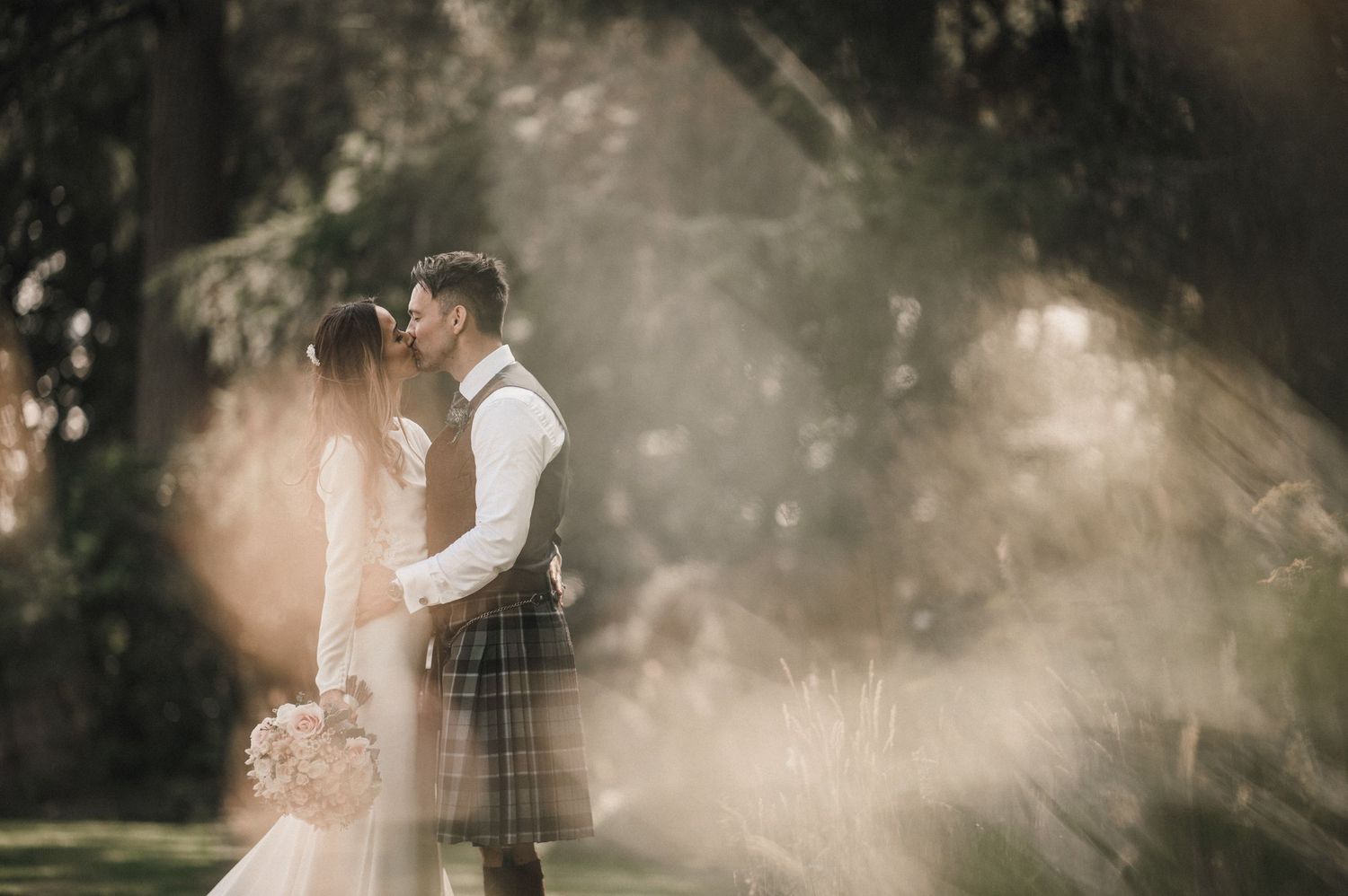 Dreamy wedding photo with light flares capturing couple in Scottish attire among forest trees.