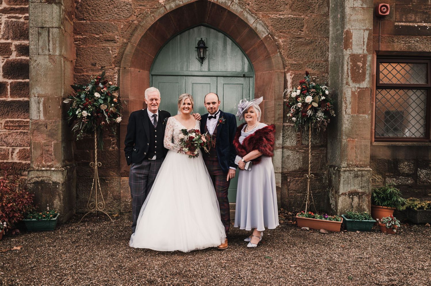 Wedding party members pose in front of blue church door with floral arrangements.