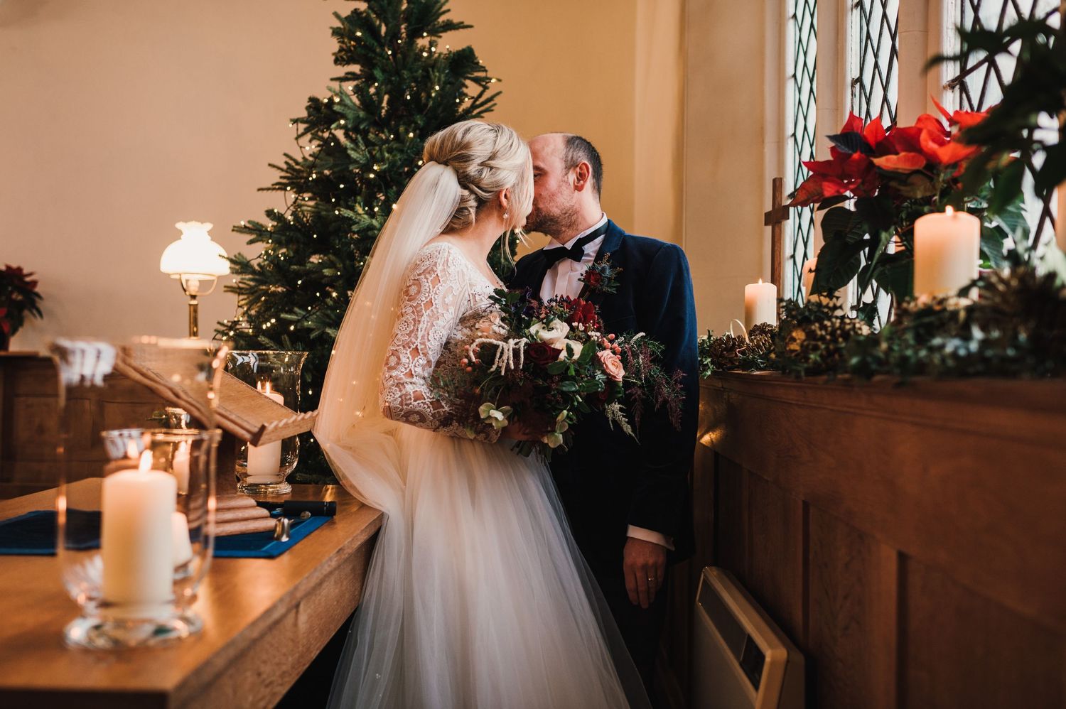 Newlyweds embrace near decorated Christmas tree and glowing candles at indoor wedding reception.