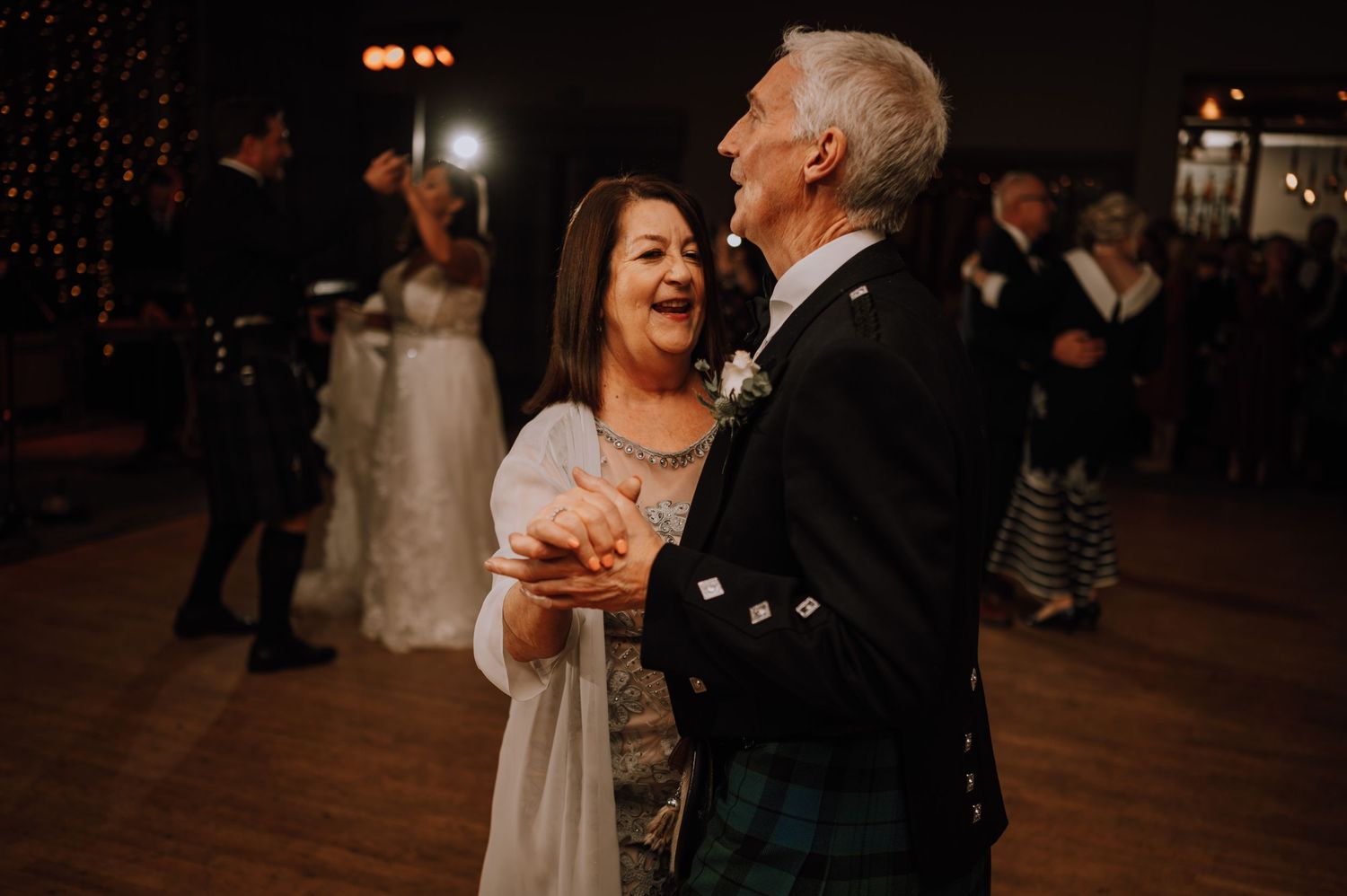 Wedding guests share dance and laughter at reception with twinkle light backdrop.