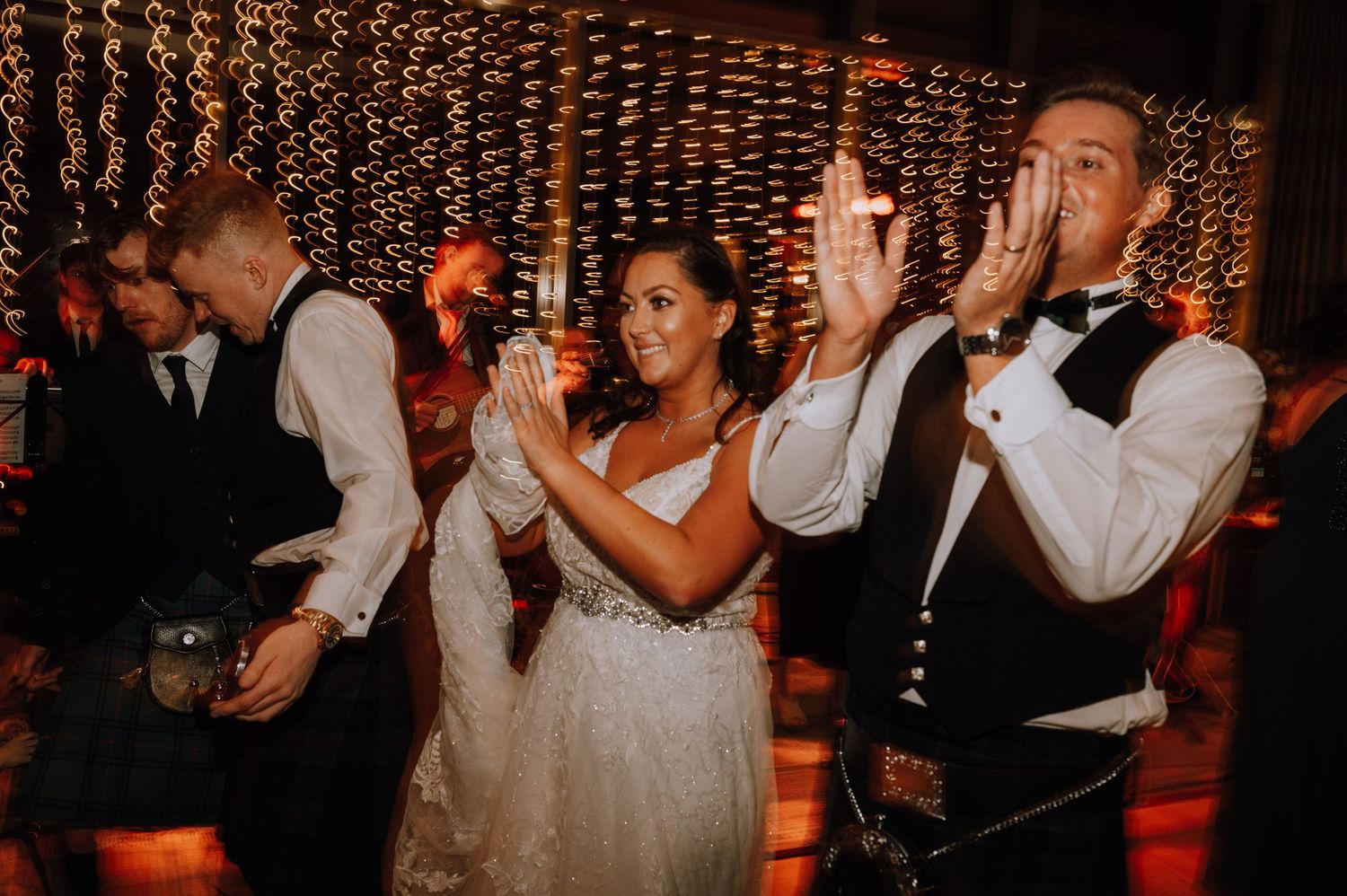 Wedding party members celebrate with raised hands under decorative string lights.