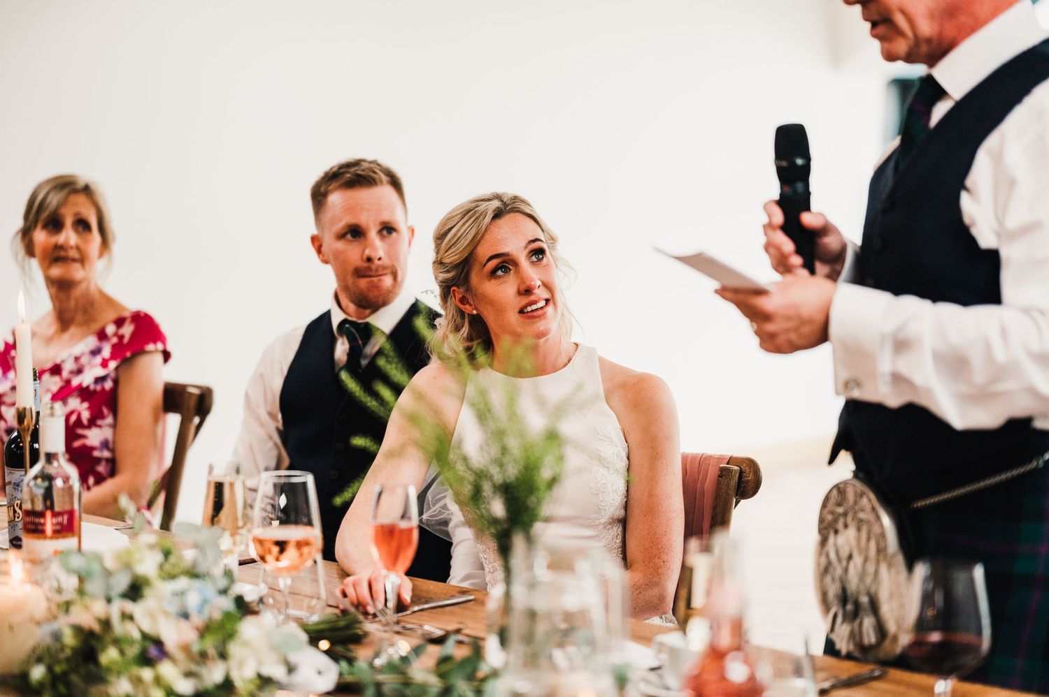 Wedding guests listen intently during a speech at an elegant reception with floral centerpieces.