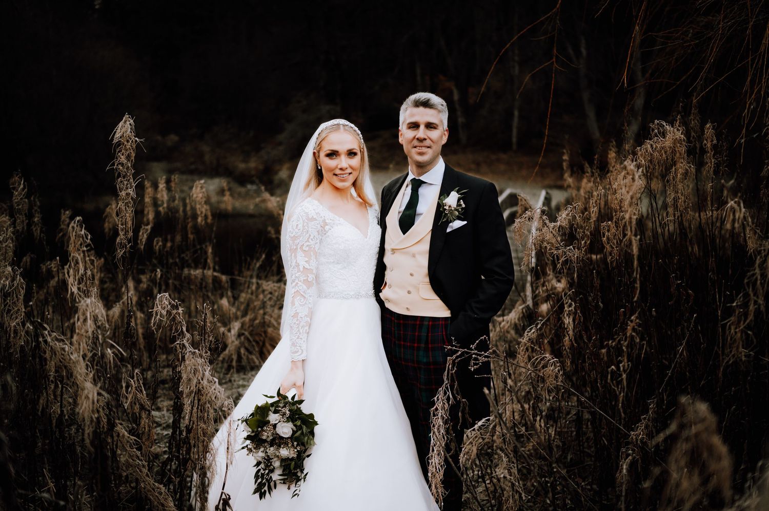 Newlyweds pose together in a rustic outdoor setting with dried grasses and dark woods.
