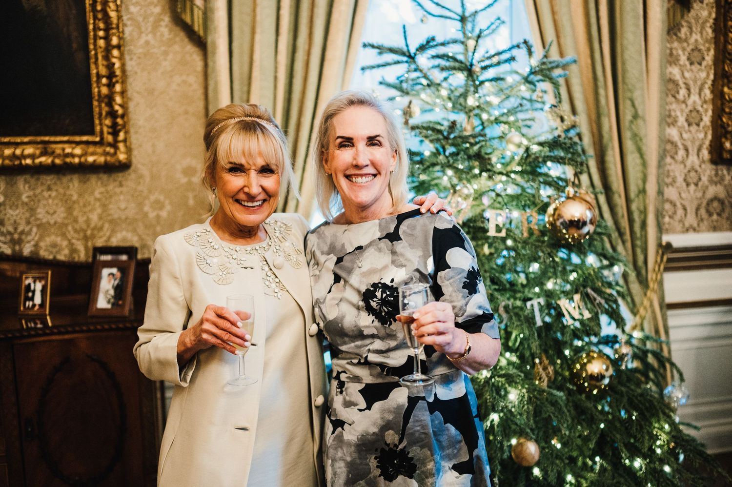 Friends gather near a decorated Christmas tree in an elegant room with ornate wallpaper.