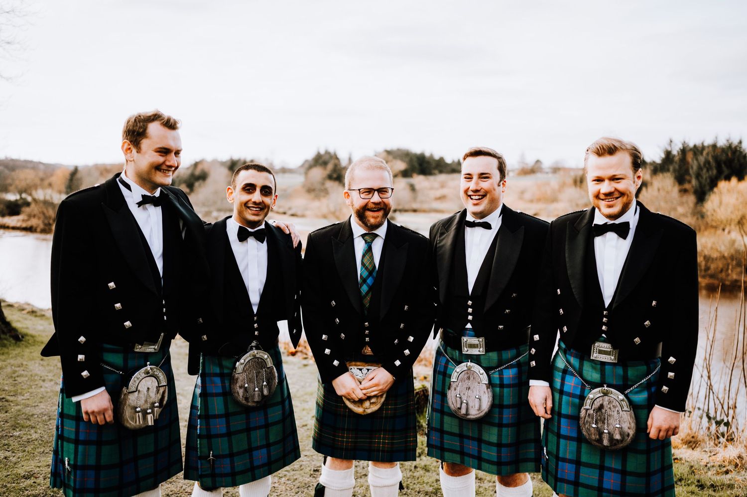 Group wearing coordinated Scottish kilts and formal wear stand together outdoors near water.