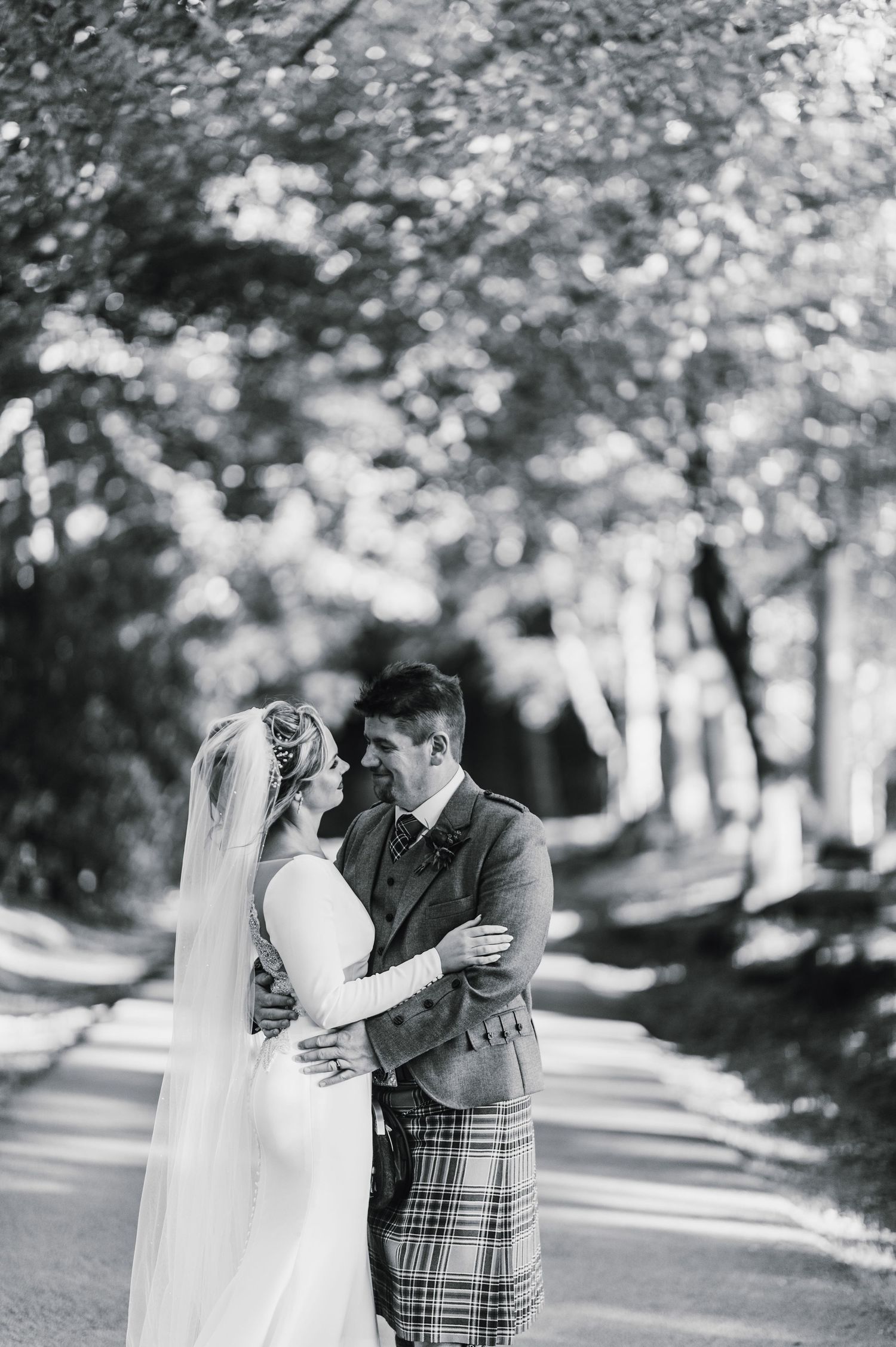 Black and white photo of newlyweds sharing an intimate moment on a tree-lined path with the groom in a kilt.