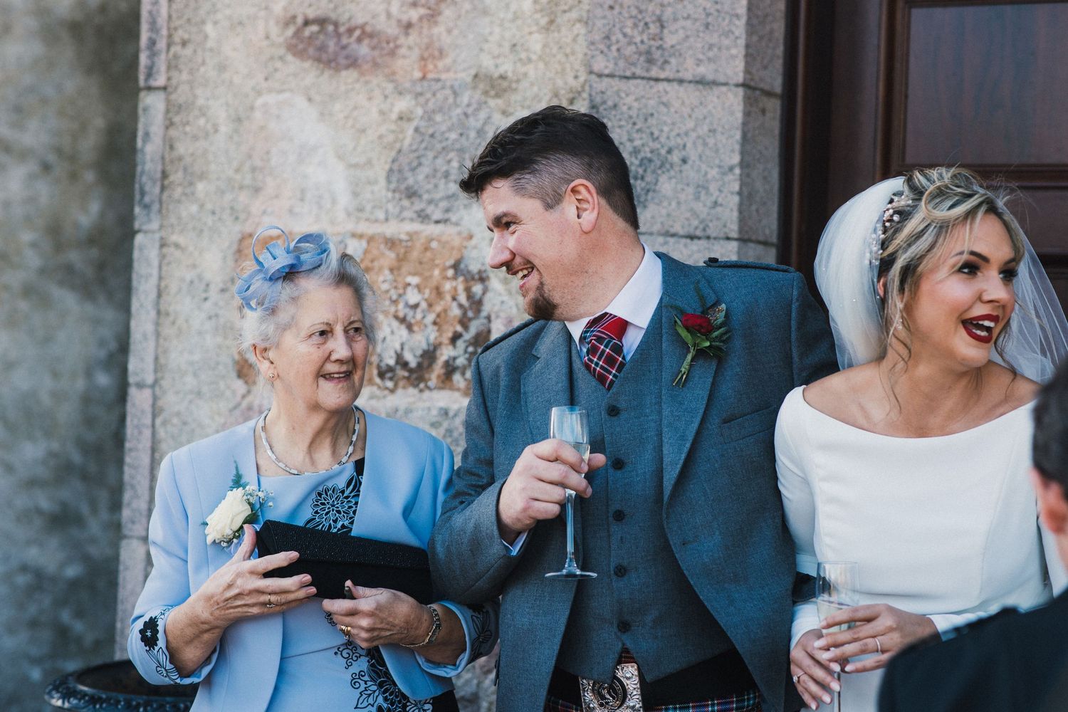 Wedding guests share a joyful moment outside a historic stone building.