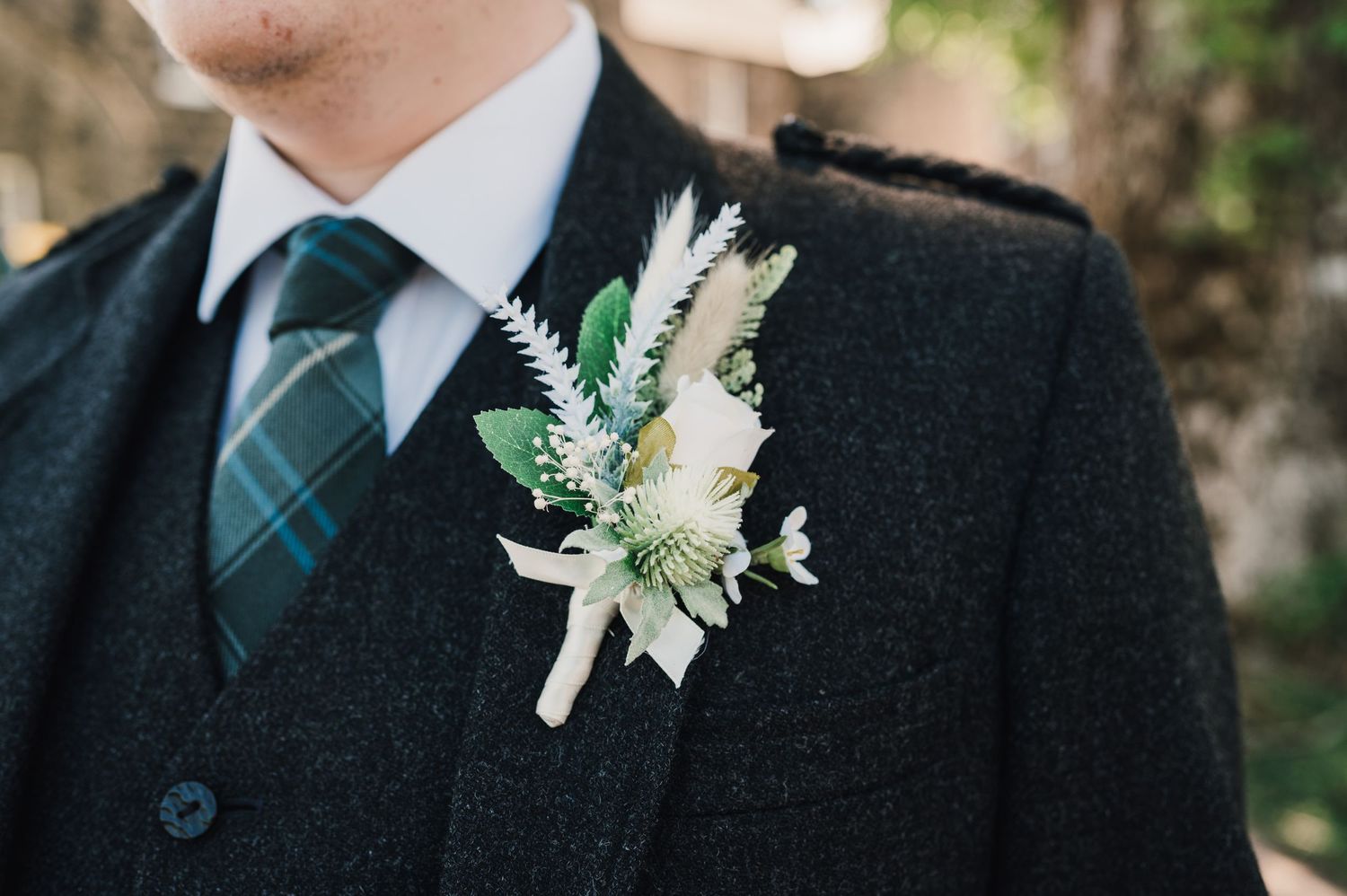 A white and green boutonnière with delicate flowers adorns a dark formal suit with tartan tie.