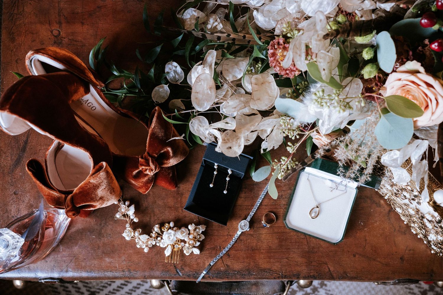 Wedding accessories including brown leather shoes, jewelry box and delicate floral arrangements on wooden surface.
