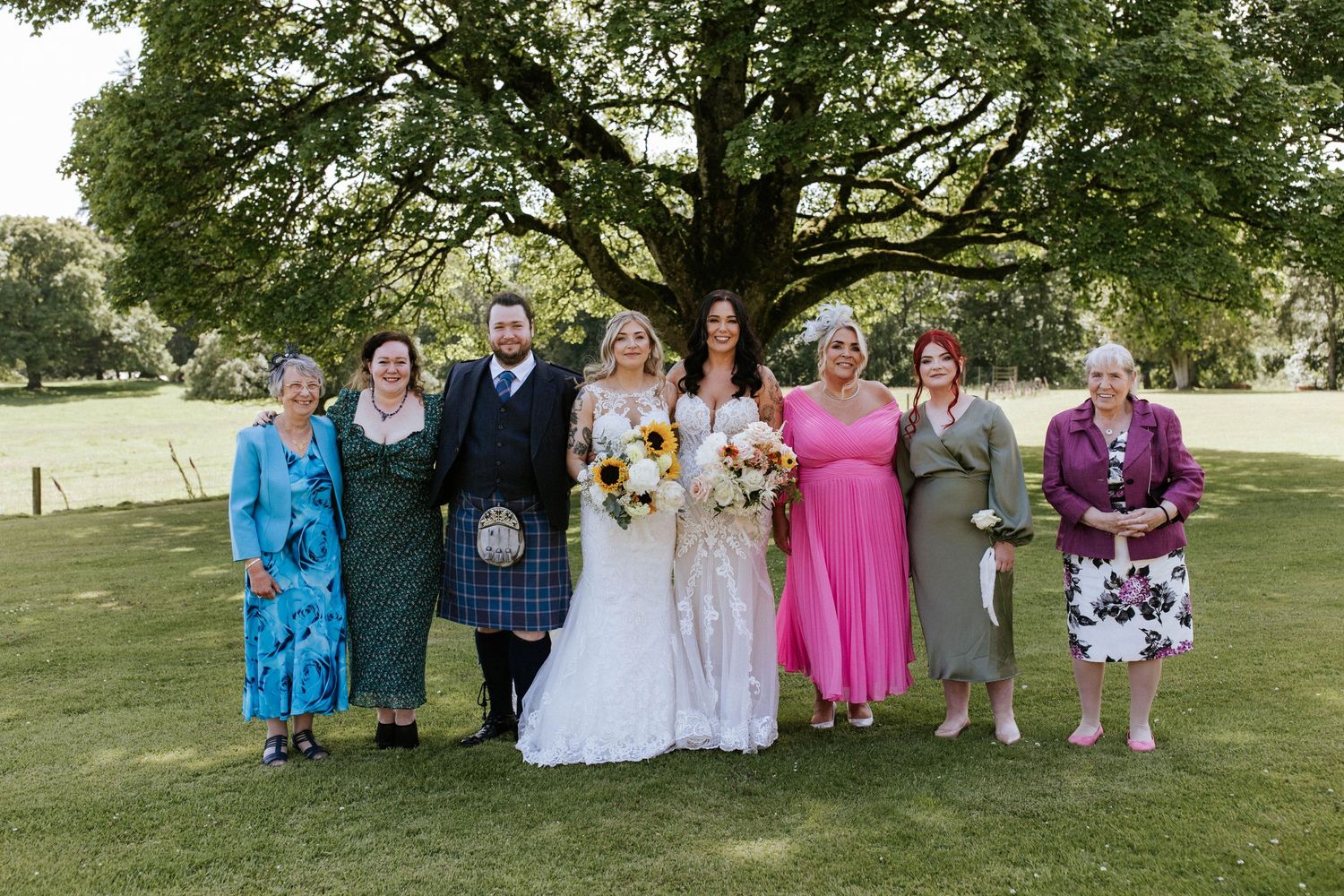 Large wedding group photo with guests in mix of formal wear and traditional Scottish attire under a tree.