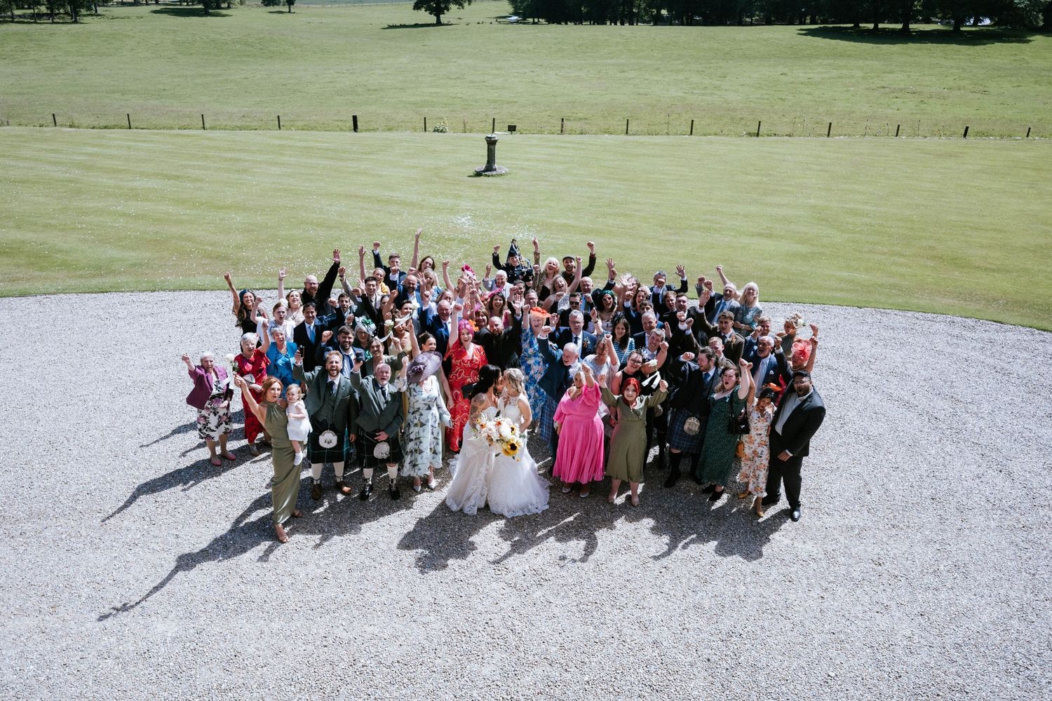 Large group photo of wedding guests posing on a grassy lawn with trees in background.