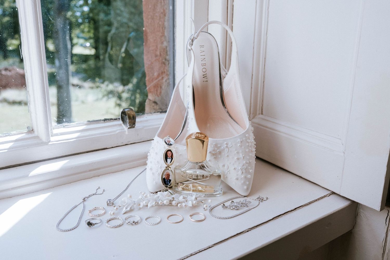 White bridal shoes with jewelry placed near a window on white surface.