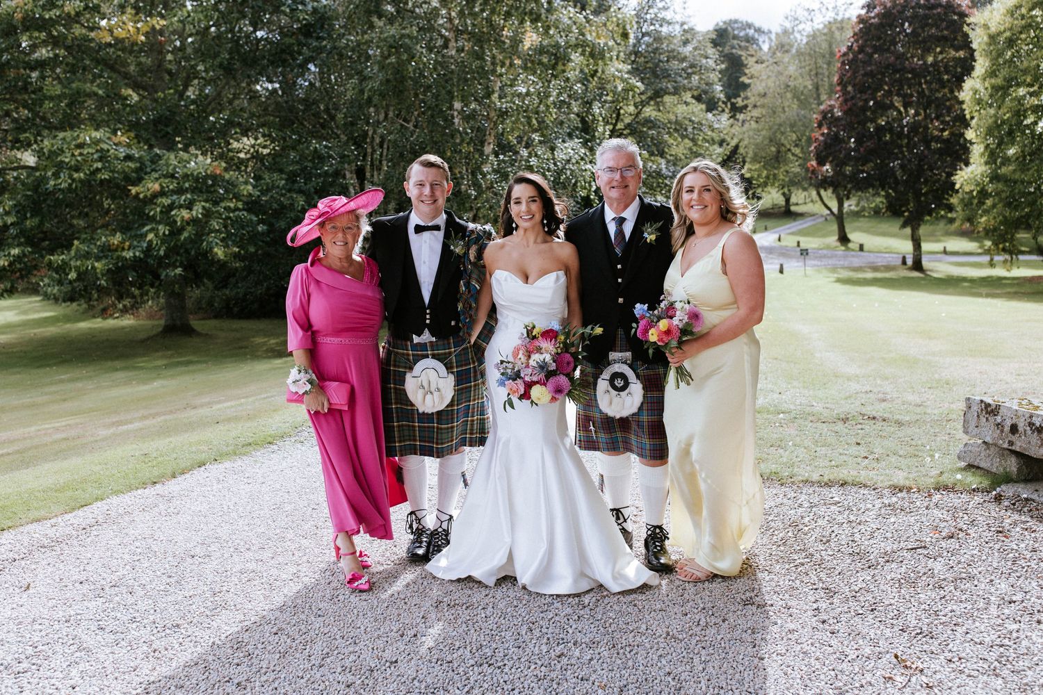 Small wedding group in mix of traditional Scottish attire and formal wear poses on garden path.