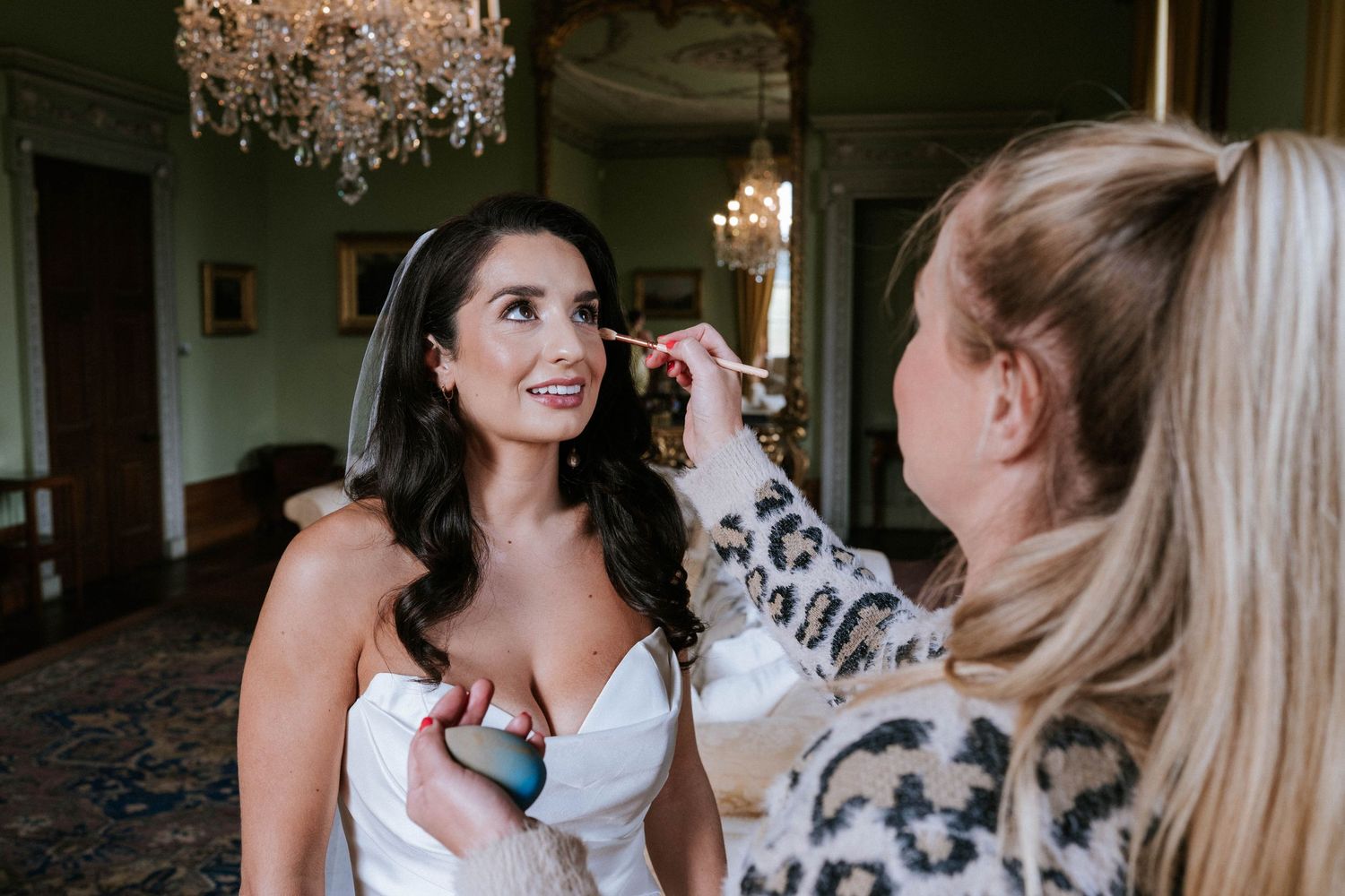 Final touches of wedding makeup being applied in an elegant room with a crystal chandelier.