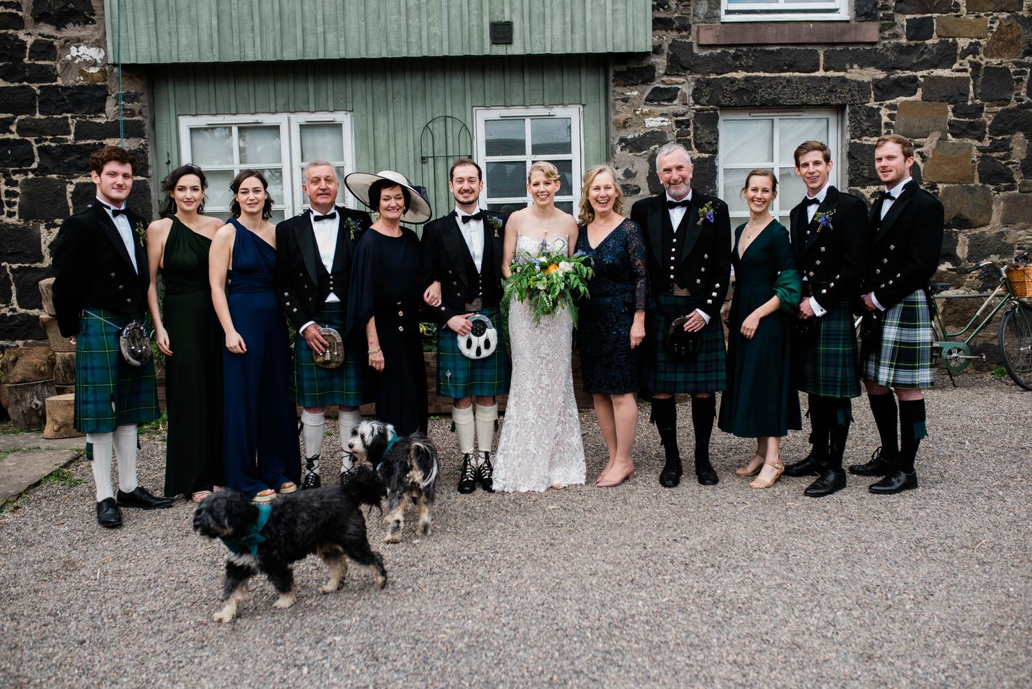 Wedding party with members in kilts and formal attire pose with a dog in front of a stone building.